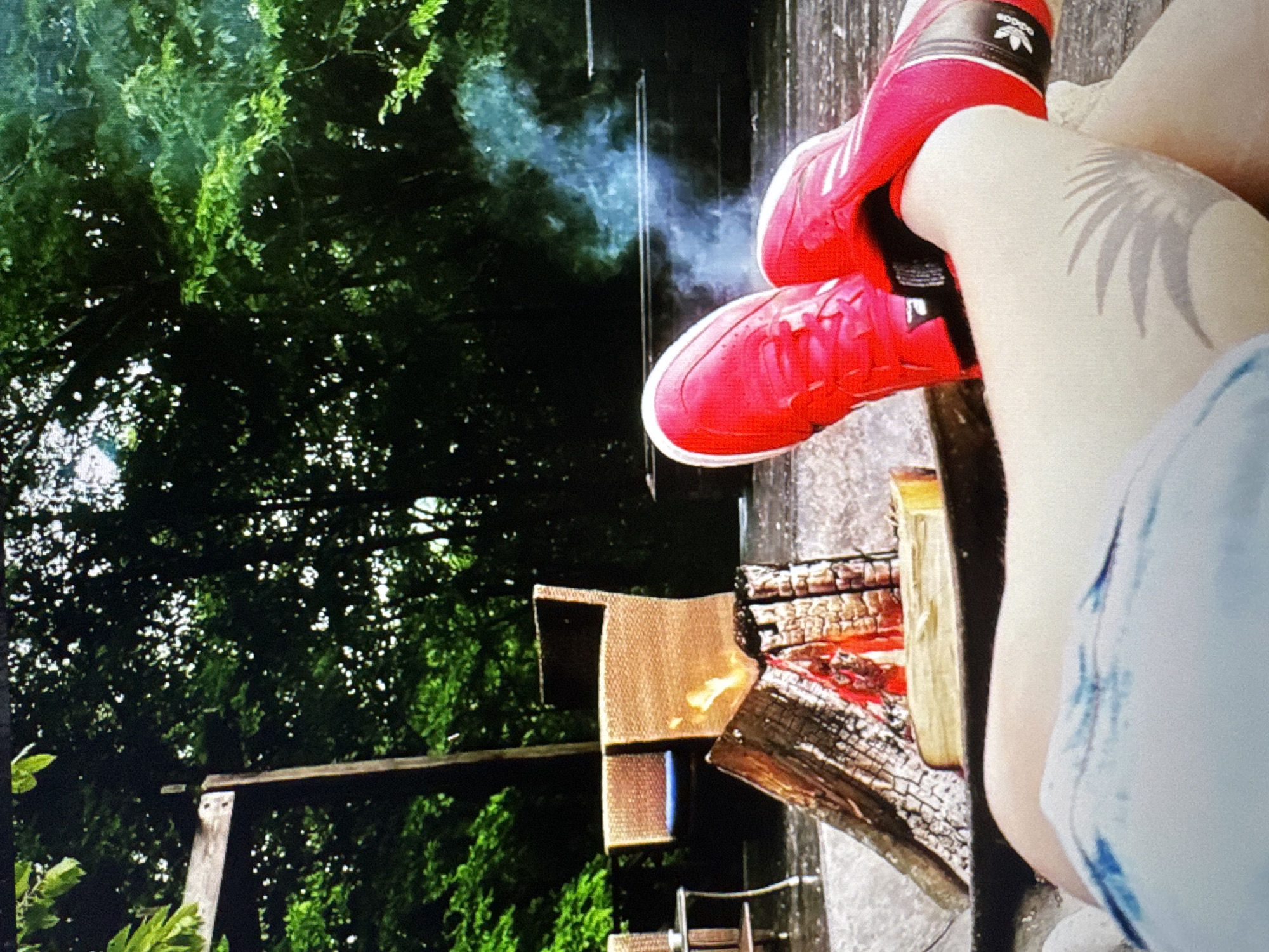 The image depicts a close-up view of a person's legs, wearing bright red athletic shoes with Adidas branding, as they relax outdoors near a fire pit. The shoes are a focal point, with the Adidas logo clearly visible on the side of the right shoe. The person is wearing light-colored denim shorts. In the background, a fire is burning in a fire pit, with logs and embers glowing. Blue-grey smoke drifts upwards from the fire. Further in the background, there are wicker chairs and a table, suggesting an outdoor seating area. Dense green foliage from trees surrounds the scene, indicating a natural or park-like setting. The lighting suggests it might be late afternoon or early evening. There is no indication of the location being in Varna, Bulgaria.