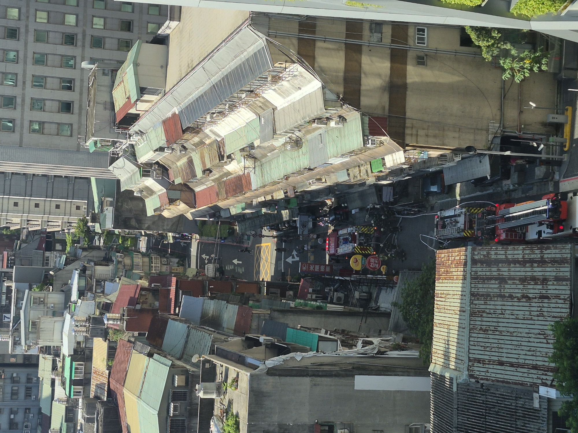 This is an aerial, daytime, medium-wide shot of a narrow street in a dense urban area, possibly Taipei, Taiwan, with many residential and commercial buildings packed closely together. A fire truck with its emergency lights on is visible in the lower center of the image, along with several firefighters in uniform surrounding it and a ladder truck behind it. The buildings are of varying heights and construction, with many featuring corrugated metal roofs in shades of green and red, as well as concrete structures. There are also some awnings and satellite dishes visible. The street itself is paved and shows traffic markings. People are visible on the street, some appearing to be involved in the emergency response. The overall impression is a busy, somewhat chaotic scene likely related to an emergency situation in a tightly packed urban environment.