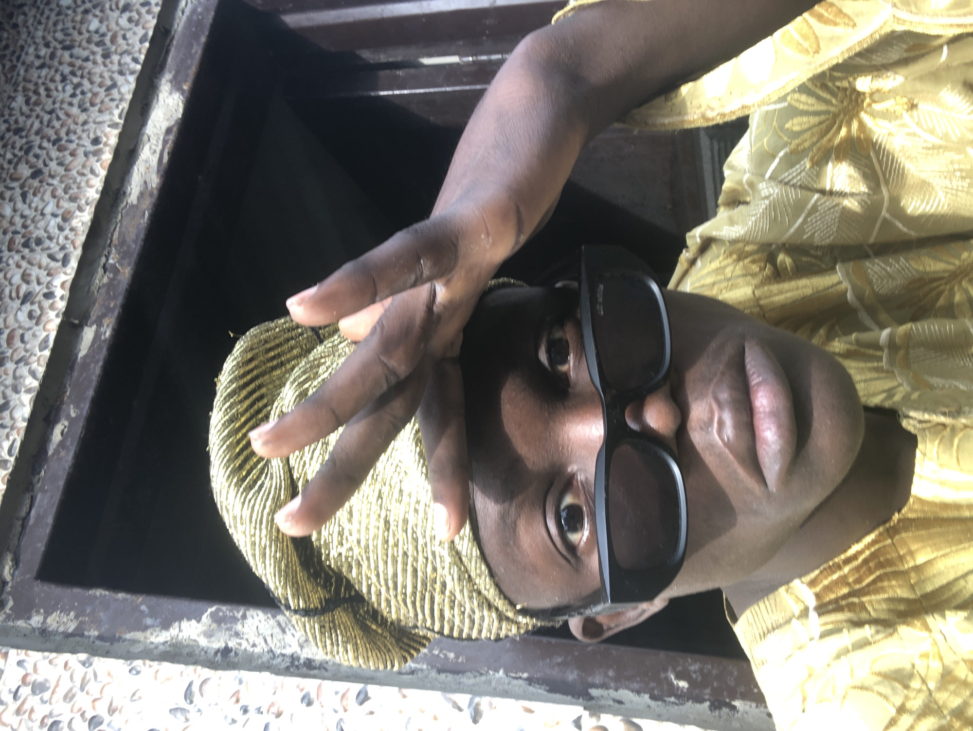 A close-up, front-facing, eye-level shot shows a dark-skinned individual wearing sunglasses, a gold-patterned headwrap, and a matching gold-patterned outfit. The individual is positioned in front of a dark opening, possibly a doorway or window, with textured, light-colored walls to the left and right. The subject's right hand is raised to their forehead, fingers splayed, with the left hand visible on the right side of the frame. The lighting suggests an outdoor setting with sunlight casting shadows on the individual's face. The image quality is moderately clear, with some graininess. The overall scene appears to be outdoors, in a somewhat rustic or weathered environment, possibly in the city of Ado, Nigeria.