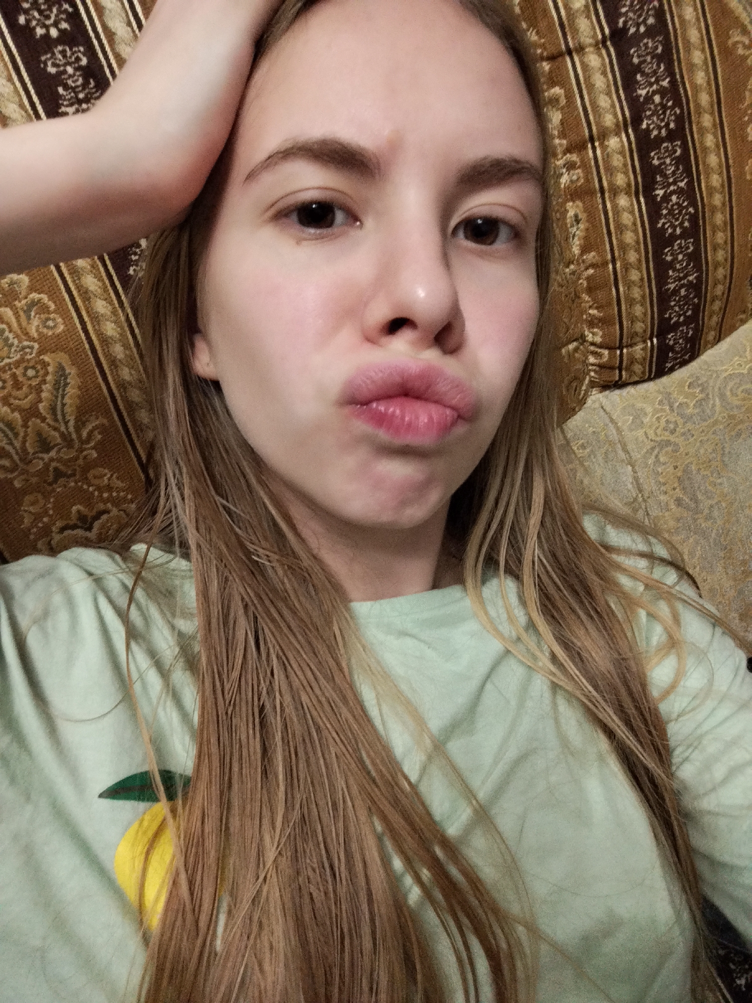 A young woman with long blonde hair is shown from the chest up, resting on a patterned sofa. She is wearing a light green t-shirt with a yellow lemon design. Her right hand is raised to her forehead, and her lips are pursed in a kiss pose. The background consists of the sofa's upholstery, which features brown and gold striped patterns. The lighting is soft and diffused, suggesting an indoor setting. The woman's expression is playful.