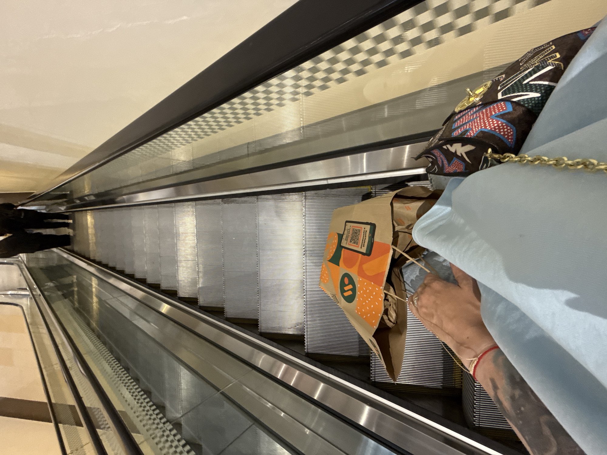 An eye-level, medium shot captures a person's perspective as they descend an escalator, holding a paper bag adorned with orange and green burger graphics, and a patterned handbag with a gold chain. The escalator steps are metallic and ribbed, leading downwards. To the left, the escalator's glass panel reflects the ribbed steps and a checkered pattern on the wall. To the right, the wall is a plain, light beige color, also featuring the checkered pattern. In the background, two individuals, viewed from the waist down, are also descending the escalator. One person wears dark pants, and the other has dark trousers and dark shoes. The overall lighting suggests an indoor environment, likely a shopping mall or similar establishment, possibly in Moscow, Russia.