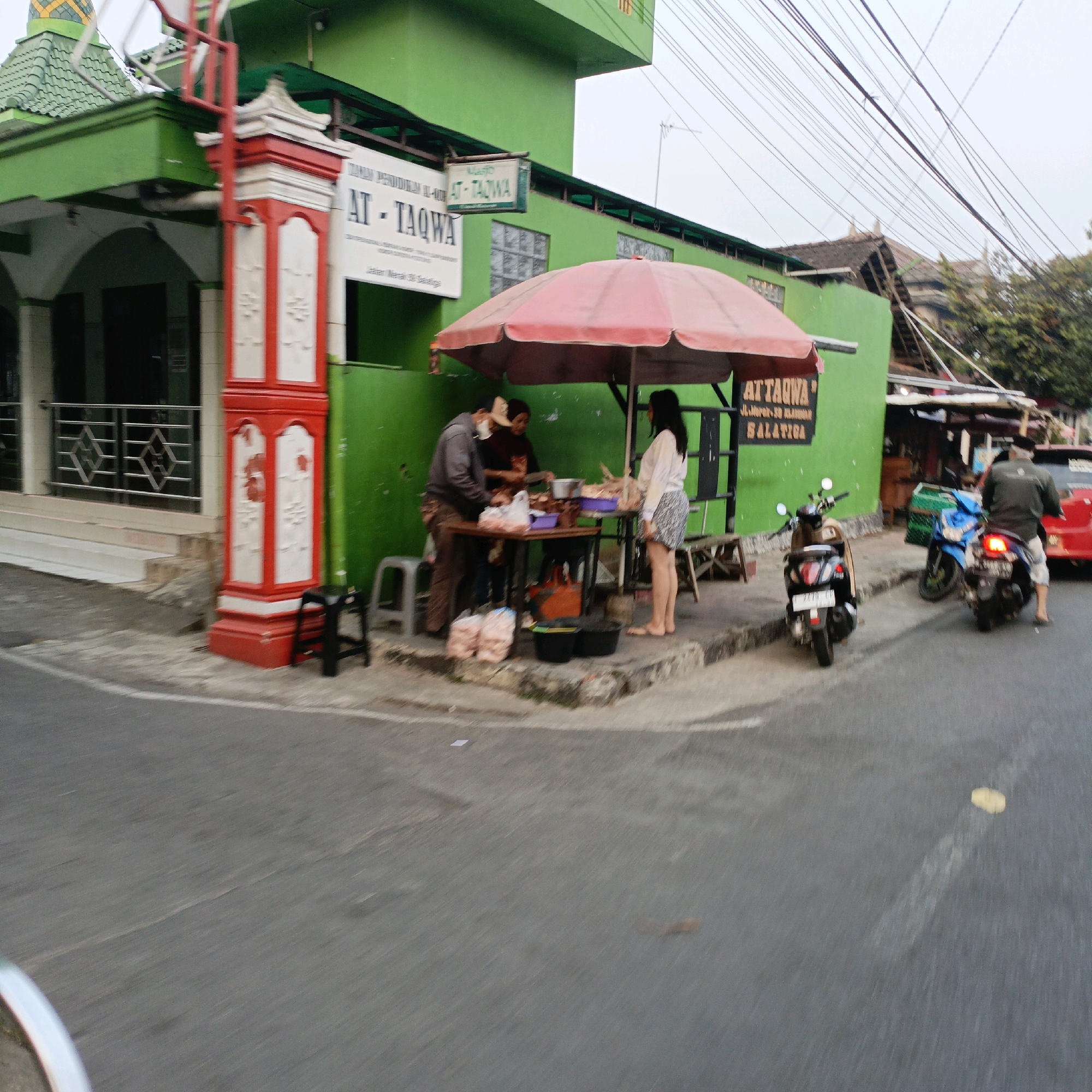 The image shows a street scene in Salatiga, Indonesia, near a green building with signs for "AT-TAQWA". In the foreground, a red and white striped pillar stands beside a road. To the right of the pillar, a woman in a white shirt and patterned skirt is standing at a table under a red umbrella, near a food stall. A man in a gray jacket and a woman in a maroon shirt are attending to the food. Several plastic bags and buckets are on the ground near the stall. On the street, a black scooter is parked. Further down the street, a blue and a black scooter are visible, with one rider on the black scooter. A red car is also present on the right side of the image. The sky is overcast.
