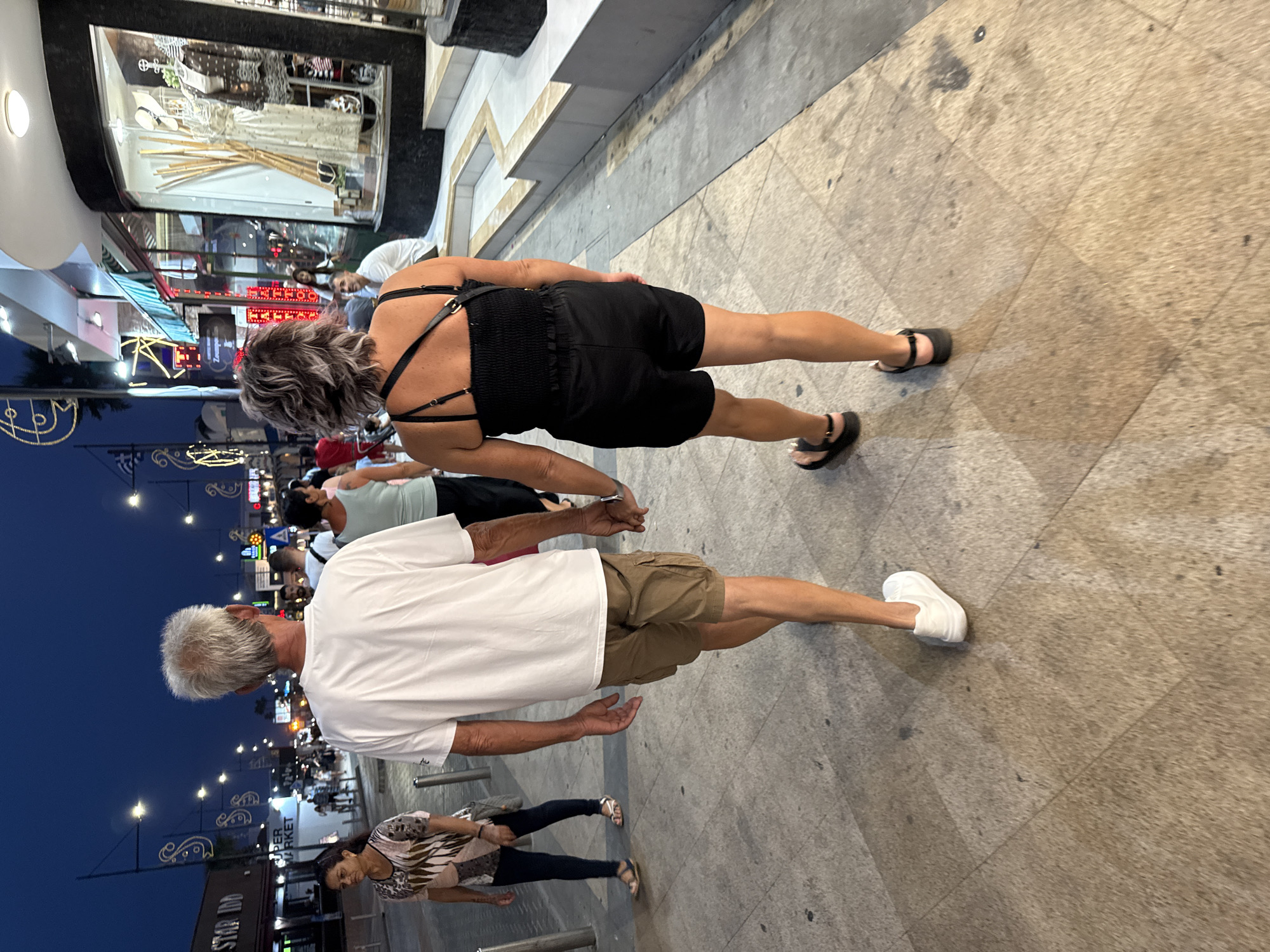 The image depicts a nighttime scene on a street in Ayia Napa, Cyprus, likely near the harbor or main strip, characterized by shops and illuminated signage. In the foreground, a couple is walking away from the camera, hand-in-hand. The man, on the left, has gray hair and is wearing a white t-shirt and khaki cargo shorts. The woman, on the right, has dark, curly hair and is wearing a black strapless top and black shorts. They are walking on a paved sidewalk with a textured, stone-like surface.

Behind them, other pedestrians are visible, including a woman in the mid-ground on the left wearing a patterned top and dark pants, and several people in the background further down the street. The street is lined with shops, one of which has a display window showcasing clothing and accessories. Streetlights and decorative lights illuminate the scene, creating a twilight atmosphere. Signs for "STAR INN" and "SUPER MARKET" are visible, as well as neon signs for businesses. The overall environment suggests a busy commercial and tourist area.