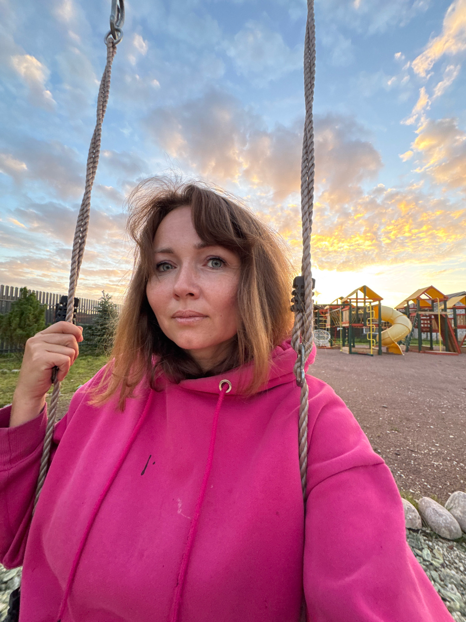 A woman with shoulder-length brown hair, wearing a bright pink hooded sweatshirt, is seated on a swing in an outdoor playground. She is holding onto the swing's rope with her left hand and looking directly at the camera. The background shows a children's playground with colorful equipment, including slides and climbing structures, set against a backdrop of a cloudy sky with hints of sunset colors. A wooden fence and some evergreen trees are visible to the left. The ground is covered in gravel. The location is identified as Kempelevo, Russia.