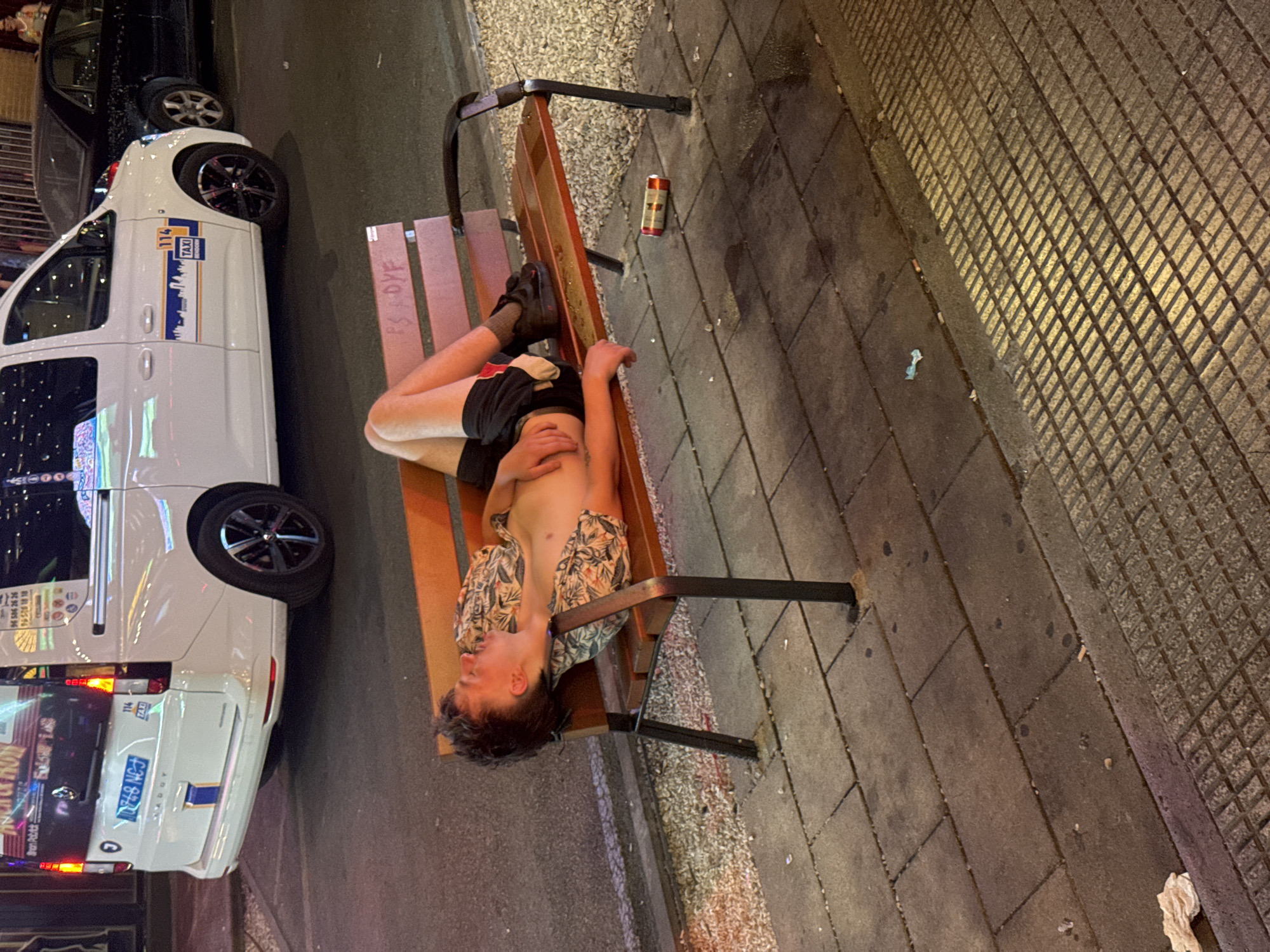 A young person, likely male, is asleep or passed out on a wooden park bench outdoors. They are lying on their back, with their head resting on their arm. They are wearing a short-sleeved patterned shirt and dark shorts. Their legs are bent, and one foot is visible. Next to the bench on the pavement is a red and gold can, possibly a beverage. The scene appears to be a street or sidewalk in a populated area, as evidenced by the paved ground, a nearby parked white van with "TAXI" branding, and a glimpse of another dark vehicle. The lighting suggests it is nighttime or late evening. The overall atmosphere suggests a sense of exhaustion or incapacitation. Text visible on the van includes "114 TAXI", a silhouette of a city skyline, and numbers "96 606 26 26" and "96518 1818". There is also some graffiti or writing on the bench itself, appearing as "PS OFF". The setting appears to be Benidorm, Spain.