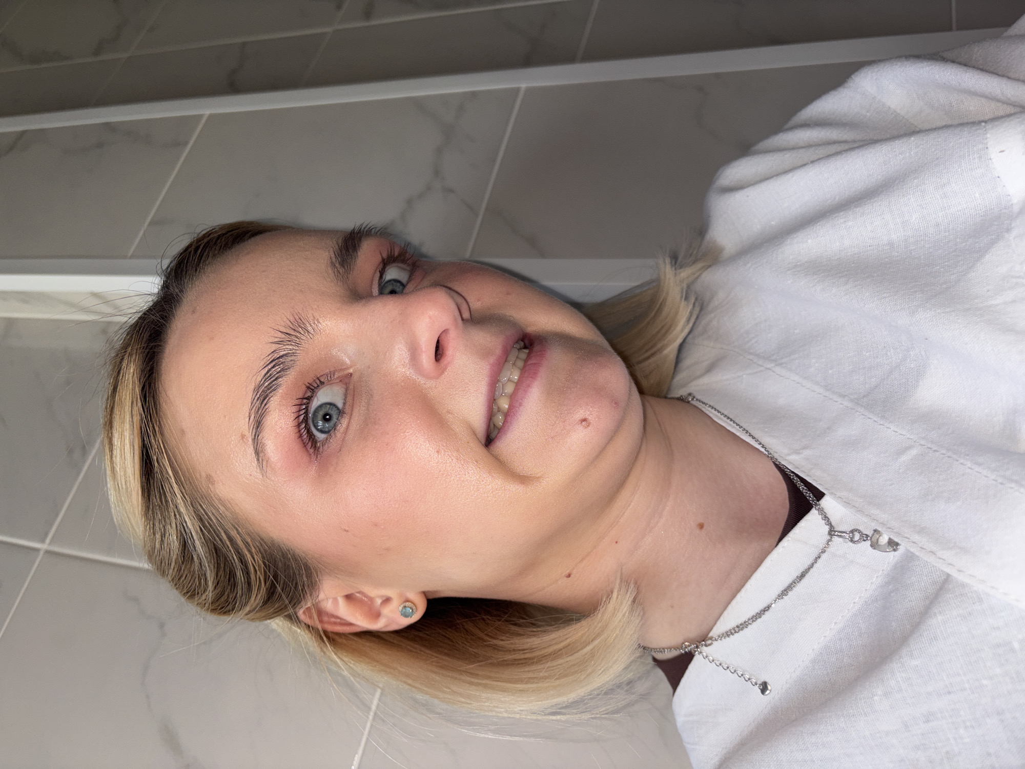 A close-up, eye-level portrait features a young woman with fair skin, bright blue eyes, and blonde hair, captured indoors in Saint Petersburg, Russia. The image is oriented with her head tilted to the right, giving the impression of looking up at the viewer. She has an alert, slightly wide-eyed expression, with her mouth slightly open in what appears to be a half-smile or a moment of surprise, revealing her upper teeth. Her eyebrows are well-groomed, and her gaze is directed straight at the camera.
She is wearing a white collared shirt, which appears to be made of a textured fabric, possibly linen. Around her neck is a delicate silver chain necklace with a small, light-colored charm that resembles a heart or a similar shape. A small, light blue stud earring is visible in her left ear. Her blonde hair is parted on the side and frames her face. Small moles are visible on her cheek and neck.
The background consists of light grey or white tiled walls or ceiling, featuring subtle patterns and straight grout lines, suggesting a modern and clean interior. The lighting is bright and even, indicating an indoor setting, likely during the daytime. No visible text is present in the image. The overall mood is direct and engaging.