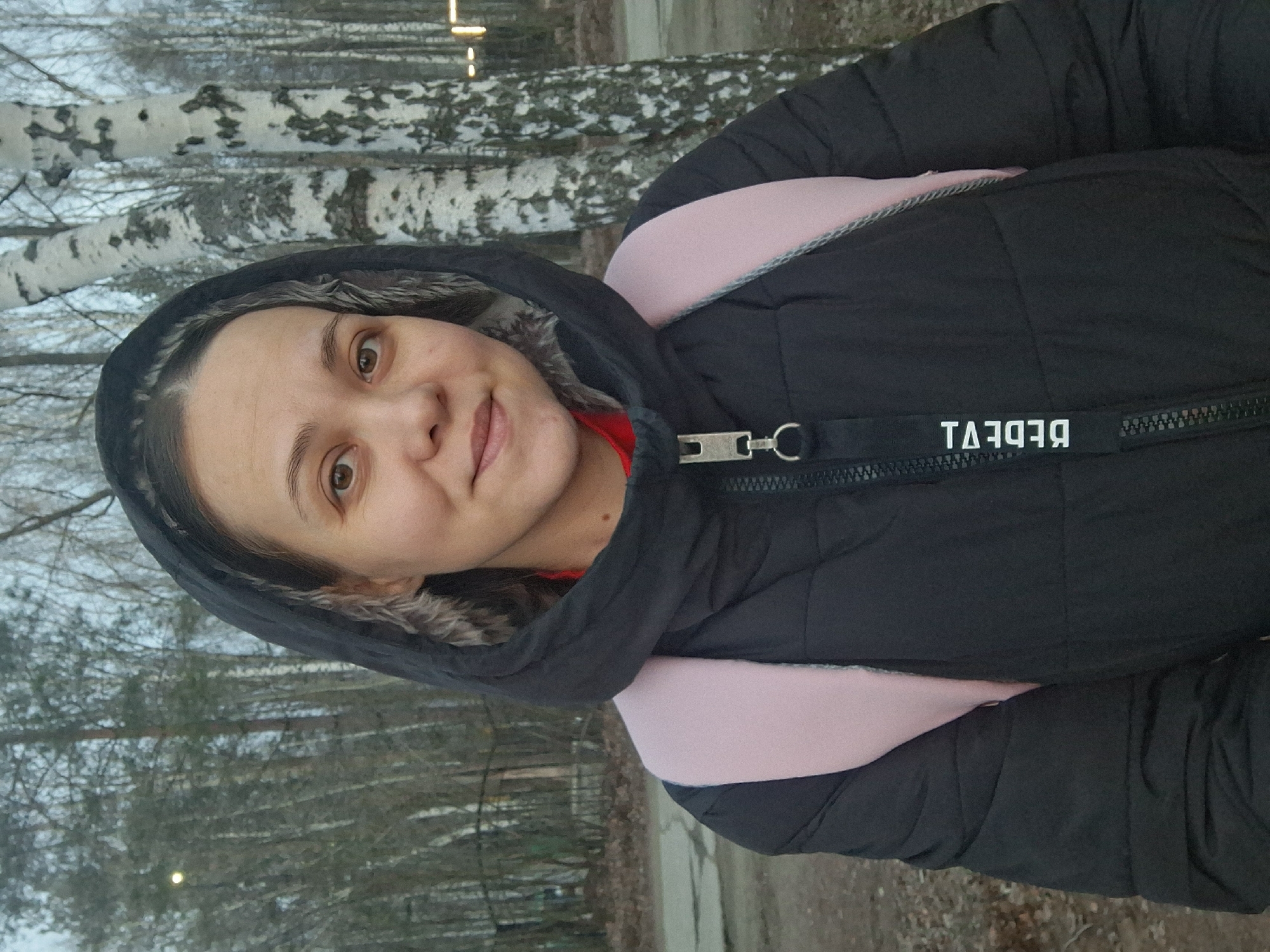 A woman with light skin, brown eyes, and dark hair is wearing a black hooded jacket over a red garment and a light pink backpack. The hood of the jacket has a fur trim. The jacket has a black tag with white lettering that reads "REBDEVI" in reverse. The woman is standing outdoors, with birch trees visible in the background. The location is Chaikovsky, Russia.