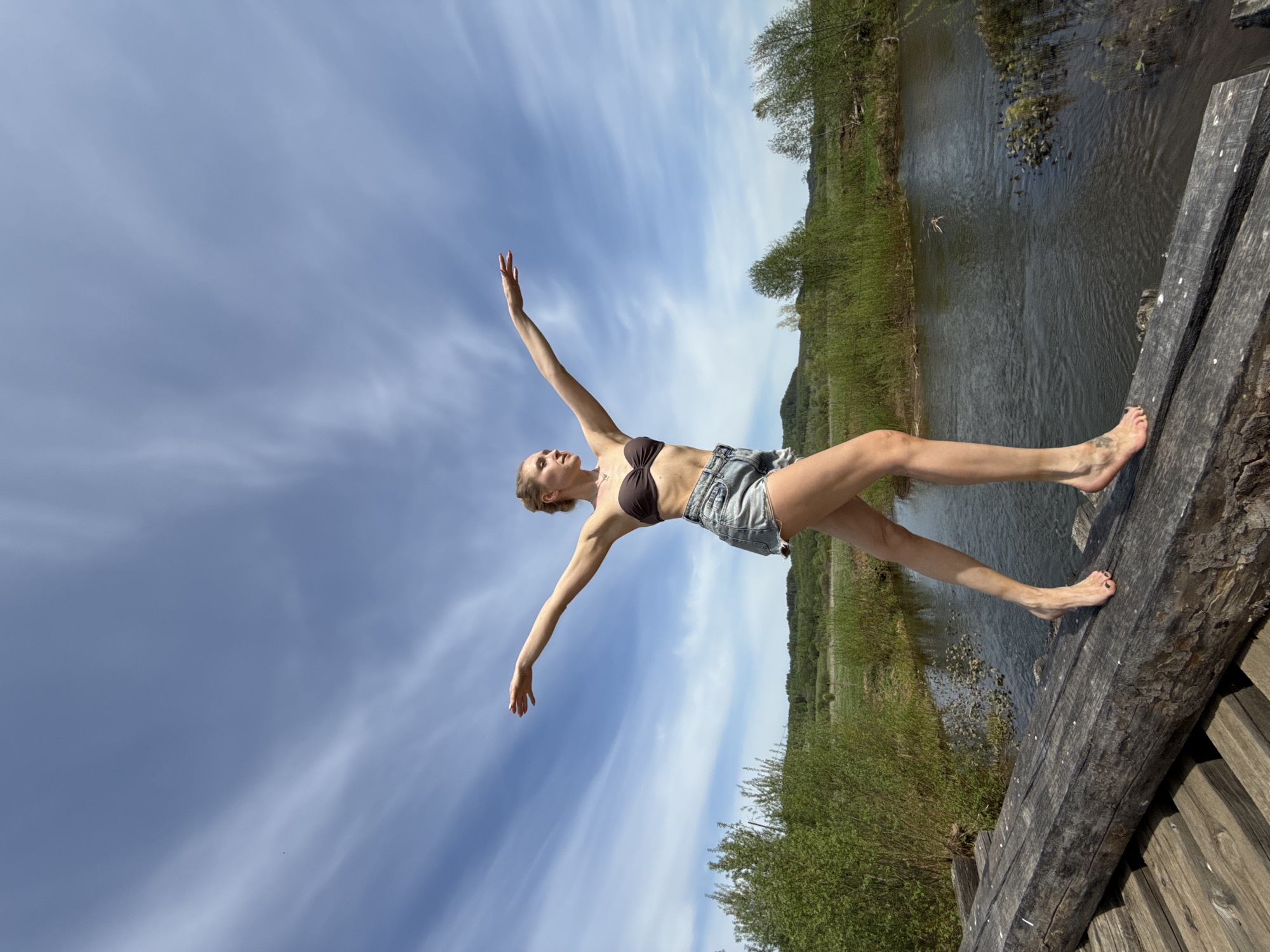 A fair-skinned woman wearing a brown bikini top and denim shorts stands on a wooden structure, possibly a bridge or platform, with her arms outstretched. She is barefoot. The structure overlooks a body of water, likely a river or stream, with green vegetation on the banks. Above, there is a bright sky with wispy clouds. The image appears to be taken in Усть-Тунтор, Russia.