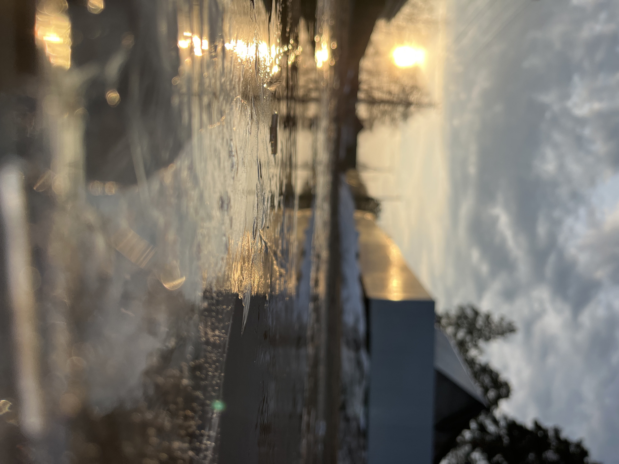 The image appears to be an abstract or distorted view of a building or monument against a sky with a bright sun or light source. The foreground shows elements that suggest a reflective surface, possibly a window or water, causing blurring and light refractions. There's a distinct architectural feature with clean lines, potentially made of metal or concrete. The sky contains clouds, and there seems to be a tree or foliage partially visible on the right. Due to the angle and abstract nature, definitively identifying the specific location in Chaikovsky, Russia, is challenging without more information.