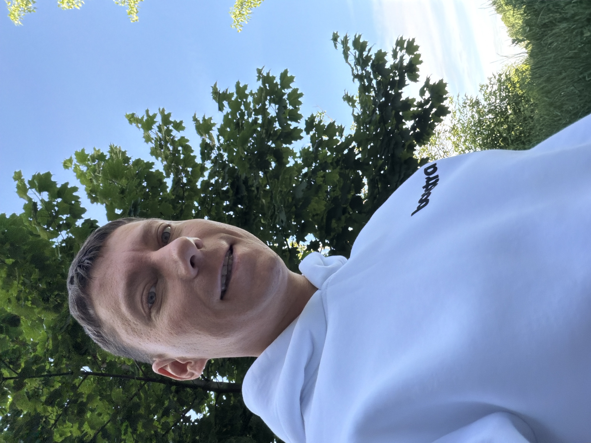 The image is a selfie taken outdoors, likely in Moscow, Russia, given the context of previous interactions. The subject is a person with short, dark hair and fair skin, looking towards the camera with a slight smile. They are wearing a white hooded sweatshirt with a black logo that appears to read "boxtra" on the left chest. The background consists of green foliage from trees and grass, with a clear blue sky overhead. The lighting suggests it is daytime, likely in the late morning or afternoon. The angle of the shot is from a low perspective, looking upwards towards the subject and the sky.