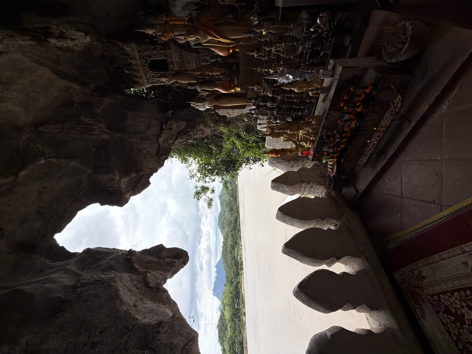 This image depicts the interior of a cave, likely a religious site, overlooking a wide, murky river. The cave entrance frames a view of lush green hills and mountains in the distance under a cloudy sky. In the foreground, within the cave, are numerous Buddhist statues of varying sizes, arranged on tiered shelves. Some of the larger statues are adorned with sashes or garlands. Offerings of flowers and lit candles are visible in front of the statues. A decorative railing with pointed, stone elements runs along the edge of the cave opening, separating the interior from the river view. A rug with an ornate pattern lies on the floor near the railing. The overall impression is one of a tranquil and spiritual sanctuary. The scene is consistent with the description of the Pak Ou Caves near Muangkeo, Laos, which are known for housing thousands of Buddha images within limestone caves along the Mekong River.