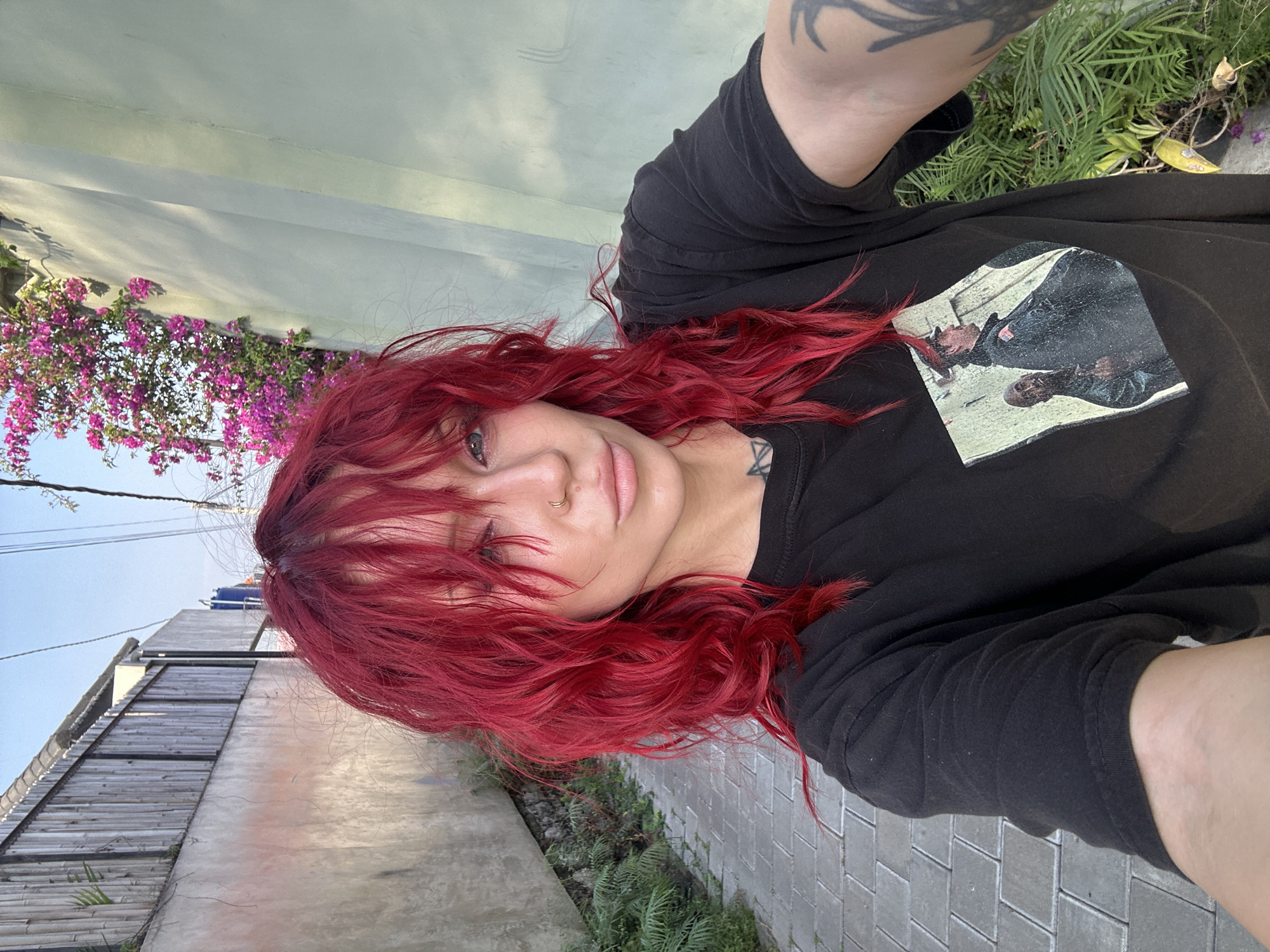 A woman with vibrant red, choppy hair and blue eyes takes a selfie. She is wearing a black t-shirt with a photo of two men printed on it. The woman has a septum ring and a tattoo on her neck. She is standing on a paved walkway in what appears to be a city alley. To her left is a textured, light-colored wall with a bamboo fence above it. To her right is a light green wall with pink flowers cascading down it. Lush green foliage is visible on the ground around her. The sky is a clear blue with some power lines visible. The scene suggests a sunny day in a tropical urban environment, possibly North Kuta, Indonesia.