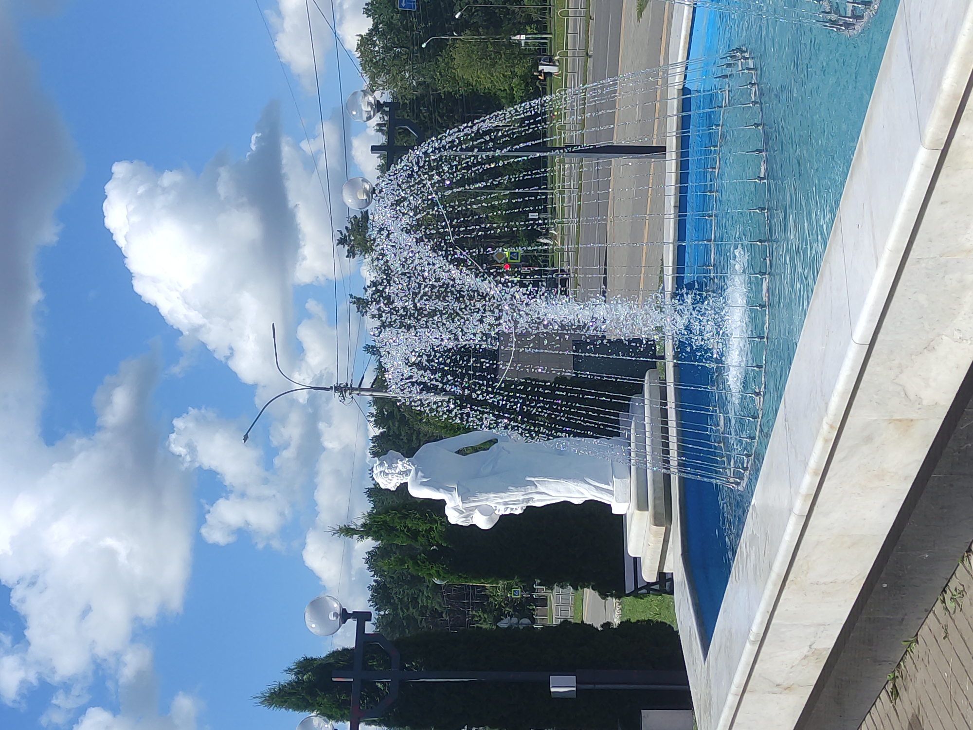 A white marble fountain with blue water is in the foreground, with water spraying upwards in a dome shape. To the left of the fountain is a white statue of a woman. Behind the fountain is a street with cars and people. A line of trees forms the background, and the sky above is blue with white clouds. The image appears to be taken in Mahilyow, Belarus, as indicated by the street signs visible in the background which are in the Cyrillic alphabet.