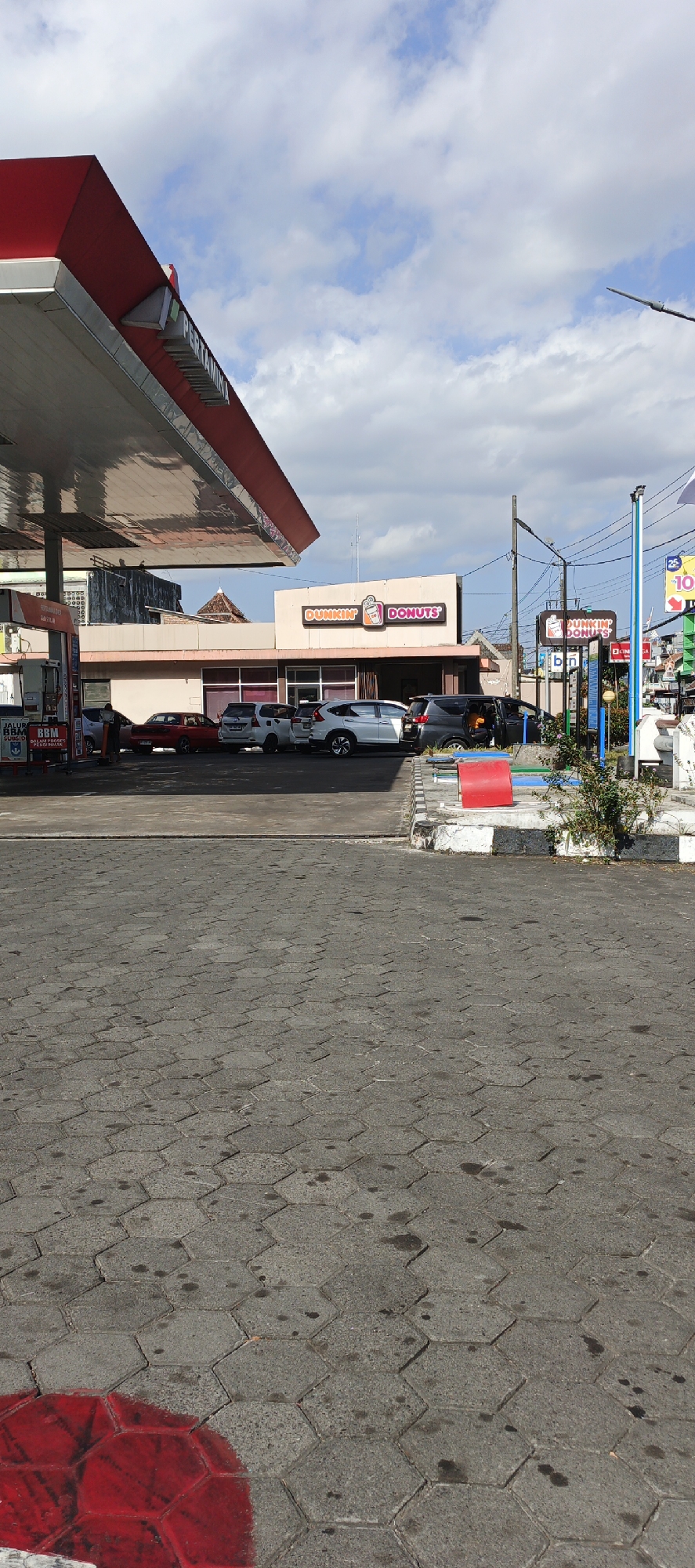The image depicts a scene at a Pertamina gas station and a Dunkin' Donuts outlet in Kasihan, Indonesia. The gas station canopy, painted red and white, extends from the left side of the frame. Several vehicles, including a red car, a white car, and a dark SUV, are parked in front of the Dunkin' Donuts building. The Dunkin' Donuts sign, featuring its iconic orange and pink logo, is prominently displayed on the building's facade. The ground is paved with hexagonal concrete blocks. The sky is partly cloudy with visible cumulus clouds. A street light pole is visible to the right of the Dunkin' Donuts building.