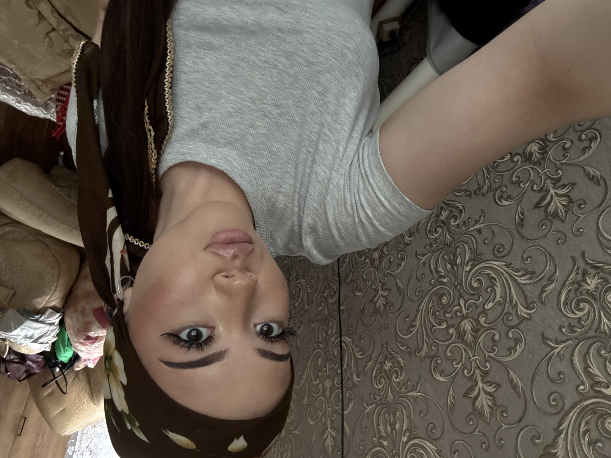 A selfie of a woman with brown hair, wearing a brown floral headscarf and a gray top, taken indoors against a wall with ornate, swirling wallpaper. The background also includes a couch or bed with miscellaneous items piled on it. It is not possible to determine whether the location is in the city of Belopakhotnyi, Russia, from the image.