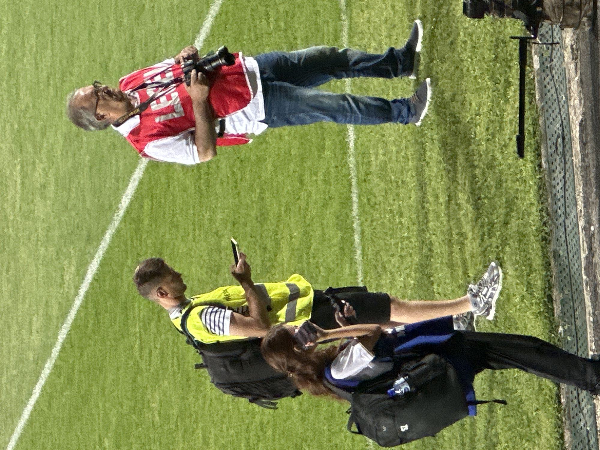 Three individuals are on a grassy field, possibly a sports stadium, in Nikšić, Montenegro.
On the right, an older man with glasses and a mustache wears a red vest over a white polo shirt and blue jeans. He holds a professional camera with a black lens in his hands. The vest has the word "PRESS" printed on it in white letters.
In the center, a younger man wearing a bright yellow vest over a striped shirt and black shorts is looking at his phone. A black backpack is slung over his left shoulder. The yellow vest also appears to have "PRESS" printed on it.
On the left, a person with dark hair, wearing a white shirt and dark pants, is holding a phone to their ear. They have a black backpack on their left shoulder and a blue strap from another bag or vest around their neck.
The ground is covered in green grass with a white line marking the edge of the field. A textured concrete border is visible in the foreground. The lighting suggests it is daytime, possibly under stadium lights.