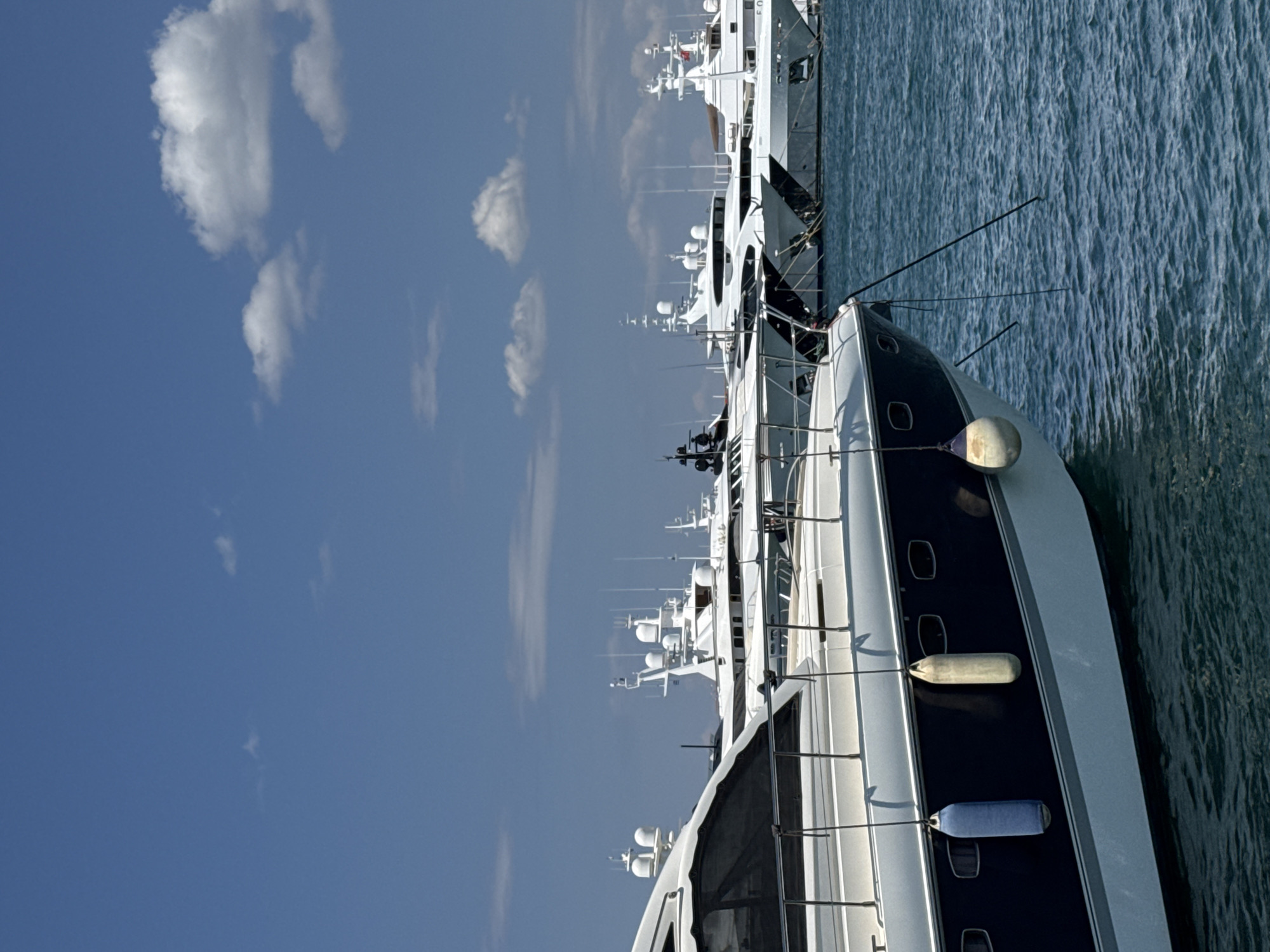 In the city of Moschato, Greece, the image captures a sun-drenched harbor scene with a clear blue sky dotted with fluffy white clouds. The foreground is dominated by the sleek, dark blue and white hull of a luxury yacht, its curved lines reflecting the bright sunlight. Two cylindrical fenders, one blue and one beige, hang from the side of the yacht, suspended by ropes. The choppy, blue water of the harbor gently laps against the boat.
In the background, a line of other large yachts and boats are docked. These vessels are predominantly white, with varying designs of superstructures and antennas reaching towards the sky. Some appear to be moored in a densely packed marina, showcasing the wealth and grandeur associated with such nautical vessels. The overall impression is one of a sophisticated maritime environment, likely a popular destination for yachting enthusiasts.
