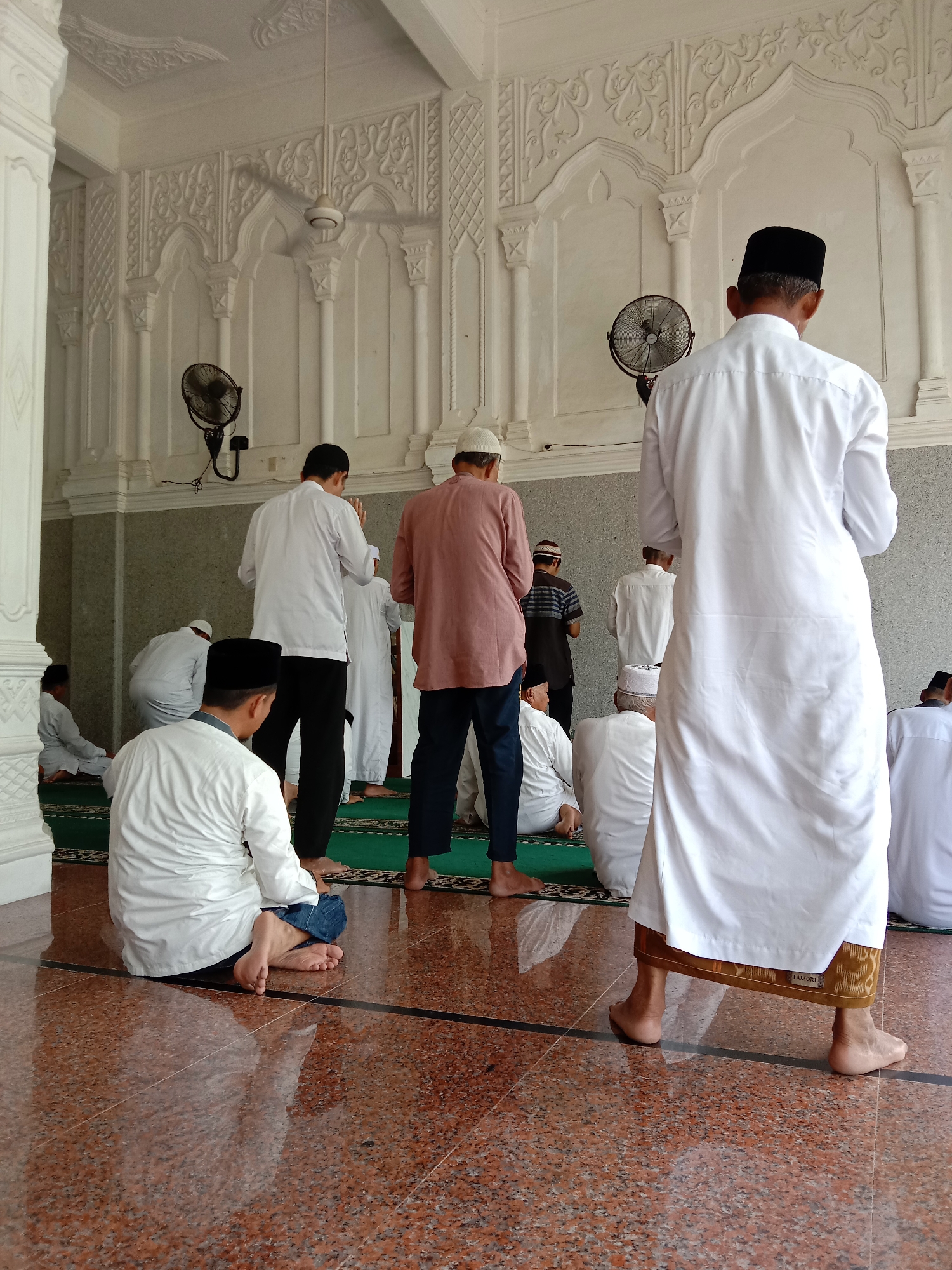 In a building that is likely a mosque, a group of Muslim men are gathered. Some are standing, and others are sitting or kneeling. All of them are barefoot and are located on a polished stone floor. A green rug covers the area where they are performing prayers. They are wearing traditional attire and head coverings. The building’s interior has white walls with ornate details, fans on the walls, and a ceiling fan. The scene is located in Lhokseumawe, Indonesia.