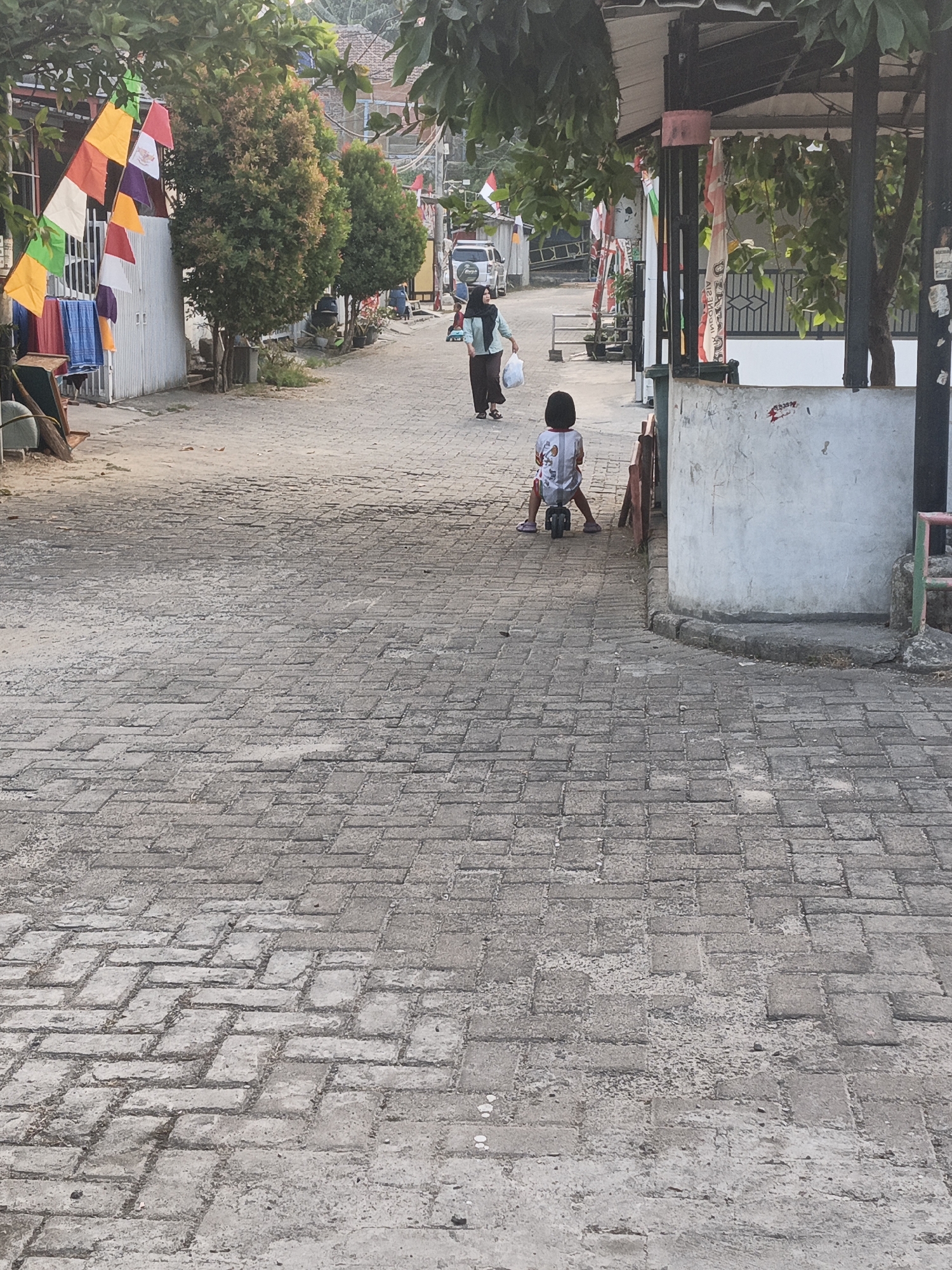 In Cilegon, Indonesia, a child is seen riding a small, three-wheeled toy bike on a paved street. The child, wearing a patterned shirt and pink shoes, faces away from the camera. A woman, dressed in a headscarf and modest clothing, walks in the background, carrying a plastic bag. The street is lined with trees and houses. Festive flags are strung across the scene, adding a touch of color. The overall environment suggests a residential neighborhood in Cilegon.