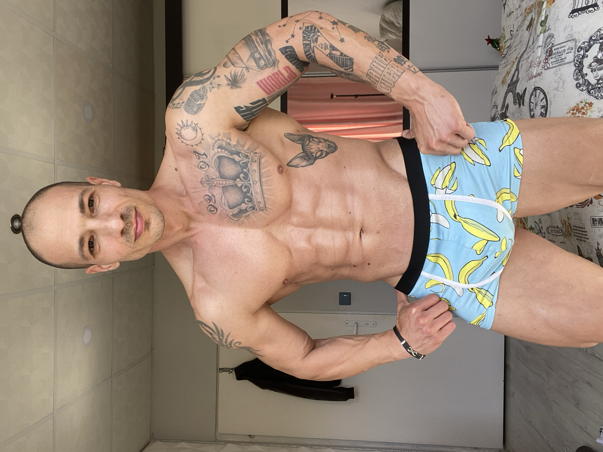 A muscular man with a shaved head stands in a room, wearing blue boxer briefs with a banana pattern. He has numerous tattoos on his arms and chest. He is holding the waistband of his underwear. The room has a light-colored wall with panels, a door, and a bed with a patterned bedspread. A black jacket hangs on a hook on the wall. The location is not specified as Pleven, Bulgaria in the image.