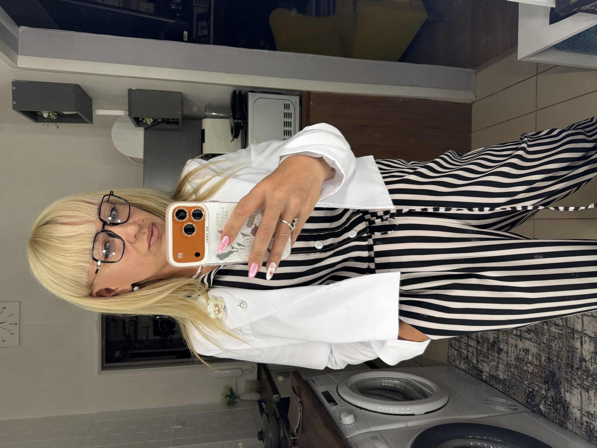 The image shows a woman taking a selfie in a bathroom setting. The woman has blonde hair and is wearing glasses, a white jacket with a flower brooch, and a black and white striped outfit. She is holding a phone with an orange phone case. The background includes a washing machine, cabinets, a mirror, and tiled walls. The specific location in the city of Chaikovsky, Russia, cannot be determined from the image.