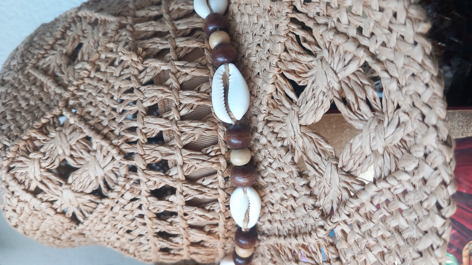 A close-up shot shows the textured woven surface of a light brown straw hat. A band adorns the hat, featuring a pattern of white cowrie shells interspersed with wooden beads of varying shades of brown and beige. The hat exhibits an open, intricate weave with floral-like patterns, revealing glimpses of a darker interior. The background is out of focus, with hints of muted colors, and the scene is not identifiable as being in the city of Άγιος Νεκτάριος, Greece.