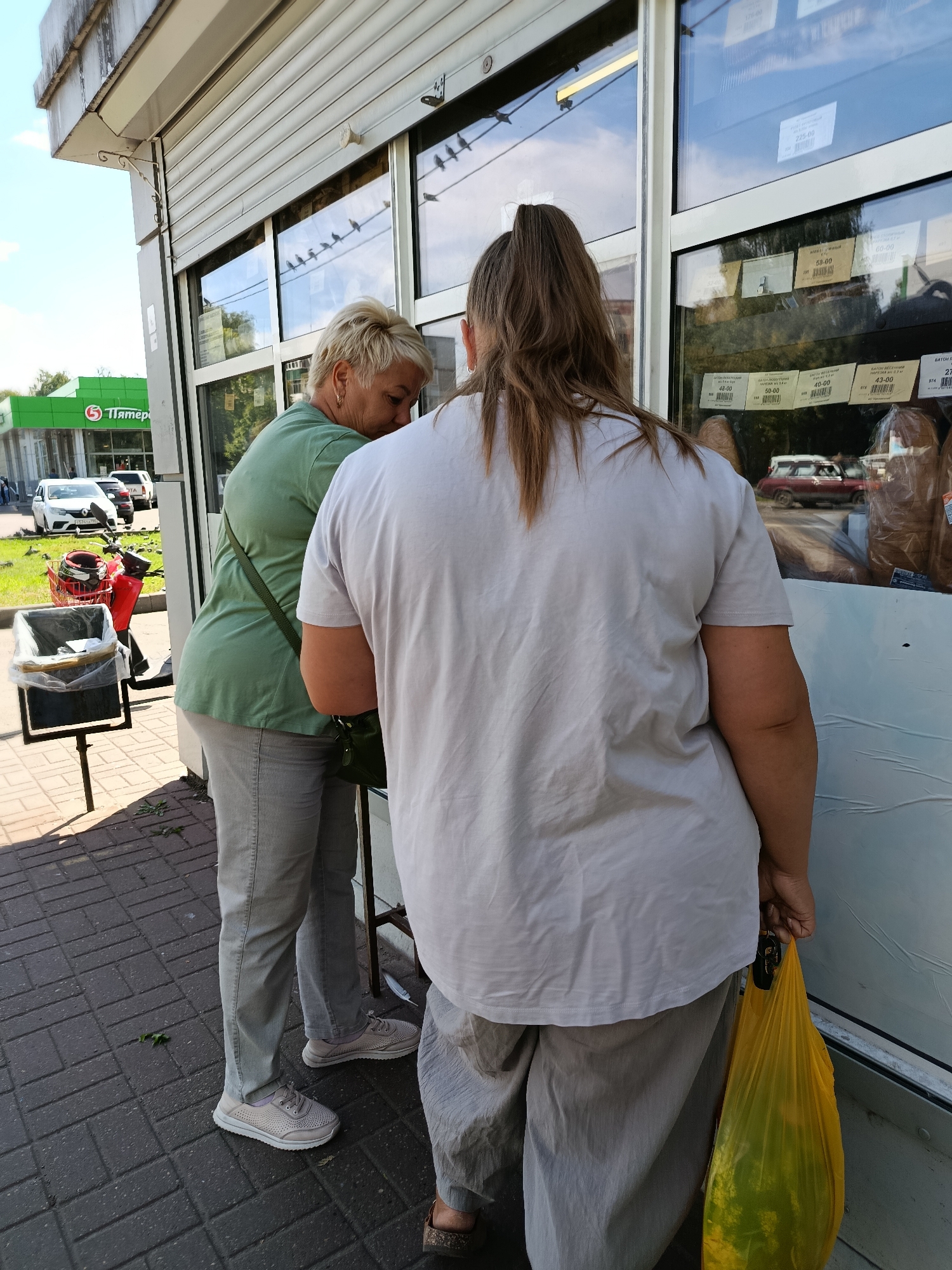 Two women are standing outside a store with large windows displaying various products. The woman on the left, with short blonde hair and wearing a green t-shirt and grey pants, is looking down. The woman on the right, with her back to the camera and long brown hair in a ponytail, is wearing a light-colored t-shirt and grey pants and is holding a yellow plastic bag. The store appears to be a food market, with price tags visible in the window. In the background, a green building with the logo "Пятёрочка" (Pyaterochka, a Russian supermarket chain) is visible. The scene is set on a paved sidewalk under a bright, sunny sky. The context suggests this is a common urban street scene in Russia, likely in a residential area or a small commercial district.