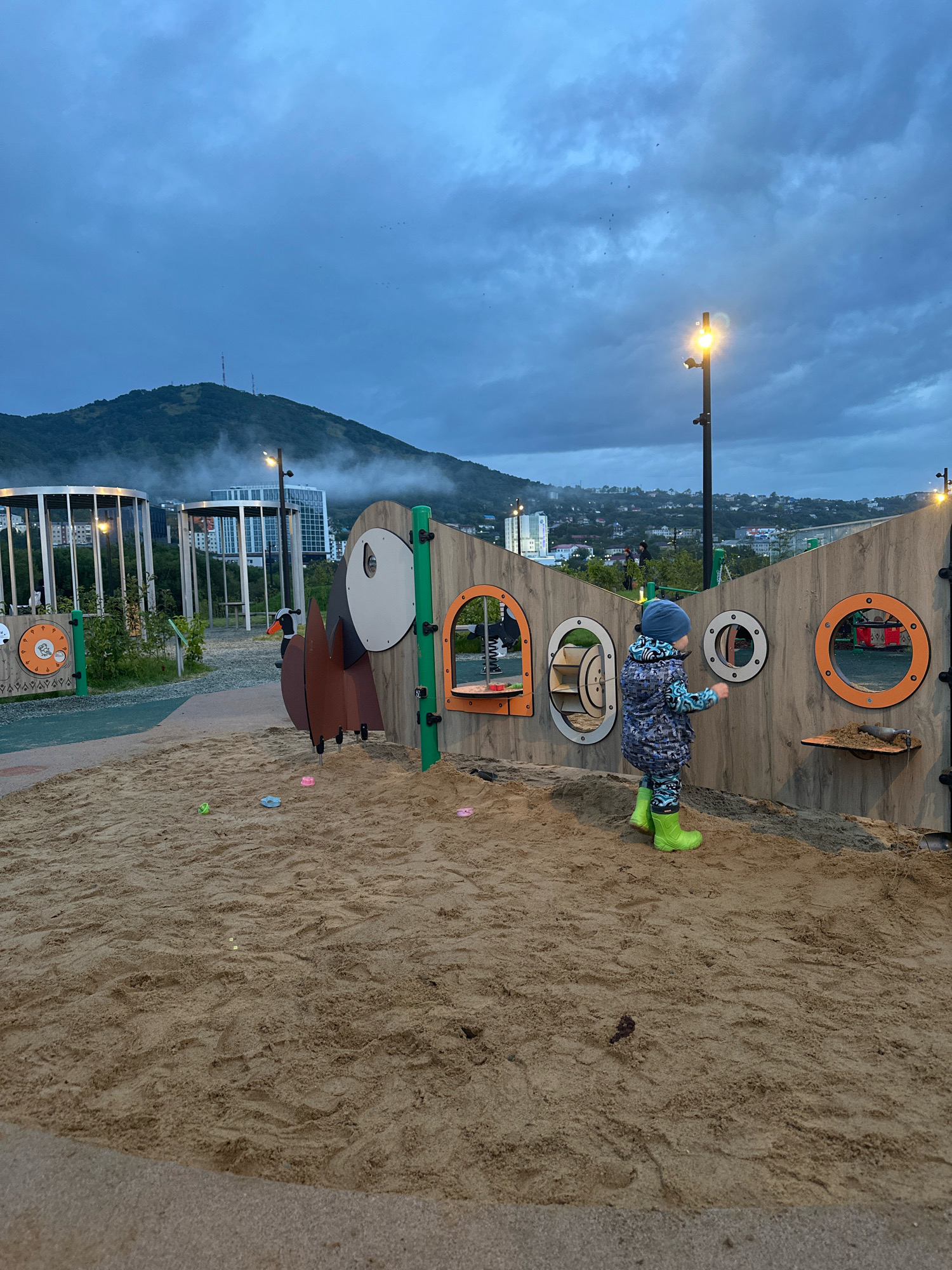 The image depicts a seaside playground in Petropavlovsk-Kamchatsky, Russia, at dusk. A young child, wearing a blue knit hat, a patterned blue and gray jacket, patterned blue leggings, and bright green rain boots, is standing in a sandpit. The child is interacting with a large wooden playground structure designed with circular openings resembling portholes. The structure is decorated with wooden cutouts of a bird. In the background, the city of Petropavlovsk-Kamchatsky is visible, with buildings nestled on a hillside under a cloudy, twilight sky. The top of a mountain, partially shrouded in mist, dominates the distant background, with two radio towers on its summit. Several lampposts illuminate the playground area, and faint outlines of other playground equipment, including circular enclosures, can be seen to the left. Small, colorful plastic toys are scattered in the sand.