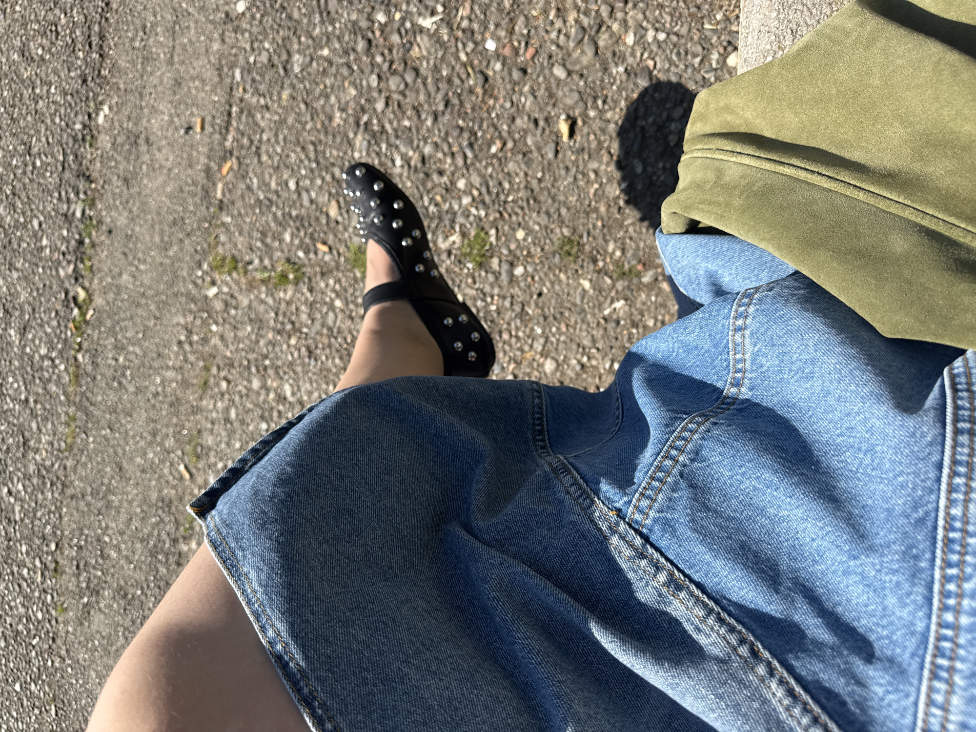 The photograph presents a downward view, showcasing a person seated outdoors. The subject wears a blue denim skirt and a green suede jacket, with a black studded shoe visible on one foot. The surface beneath consists of asphalt and stone. There is no evidence to confirm this image was taken in Milan, Italy.