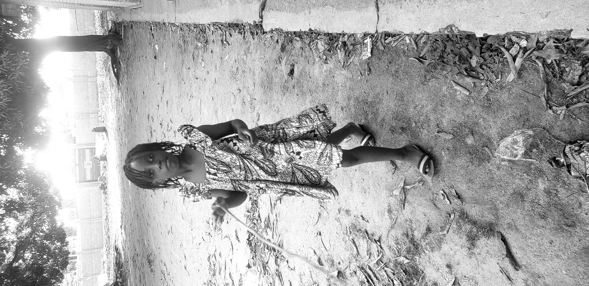 A young Black girl stands on a dirt path in Narayi, Nigeria, holding a skipping rope. She is wearing a patterned dress and sandals. The sun is setting, casting long shadows and creating a bright glare in the background. The environment appears somewhat neglected, with scattered leaves and debris on the ground. A tree is visible to the right of the girl, and a wall and fence can be seen in the distance.