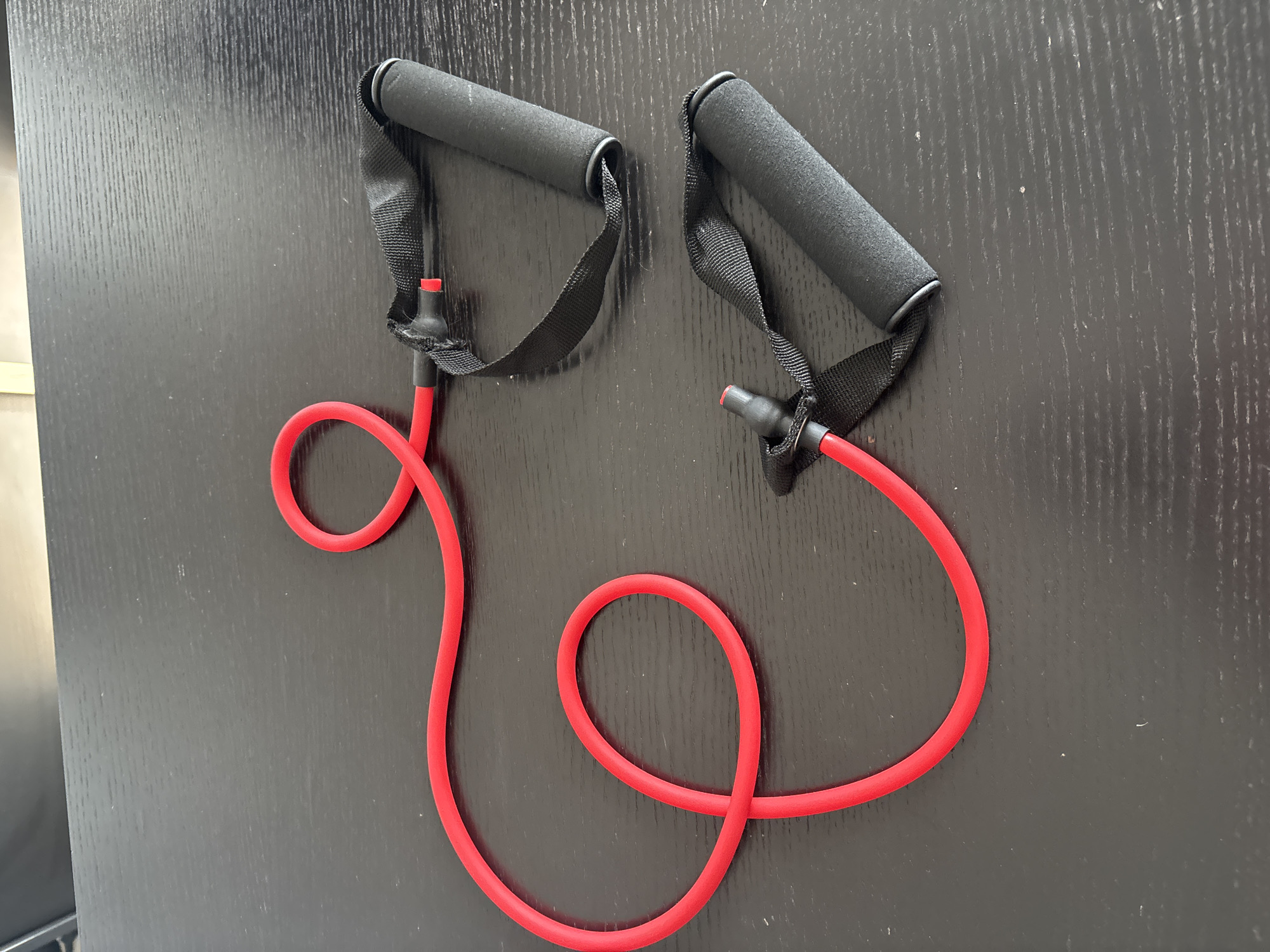 This is a top-down, eye-level, medium shot of two red resistance bands with black foam handles lying on a dark, textured surface. The bands are positioned diagonally across the frame, with the top band slightly above and to the right of the bottom band. Both bands are coiled, forming loops. The handles are cylindrical and appear to be made of foam for grip. The surface is a dark, wood-like material with a visible grain. There is no indication of the city of Dubai, United Arab Emirates in the image.