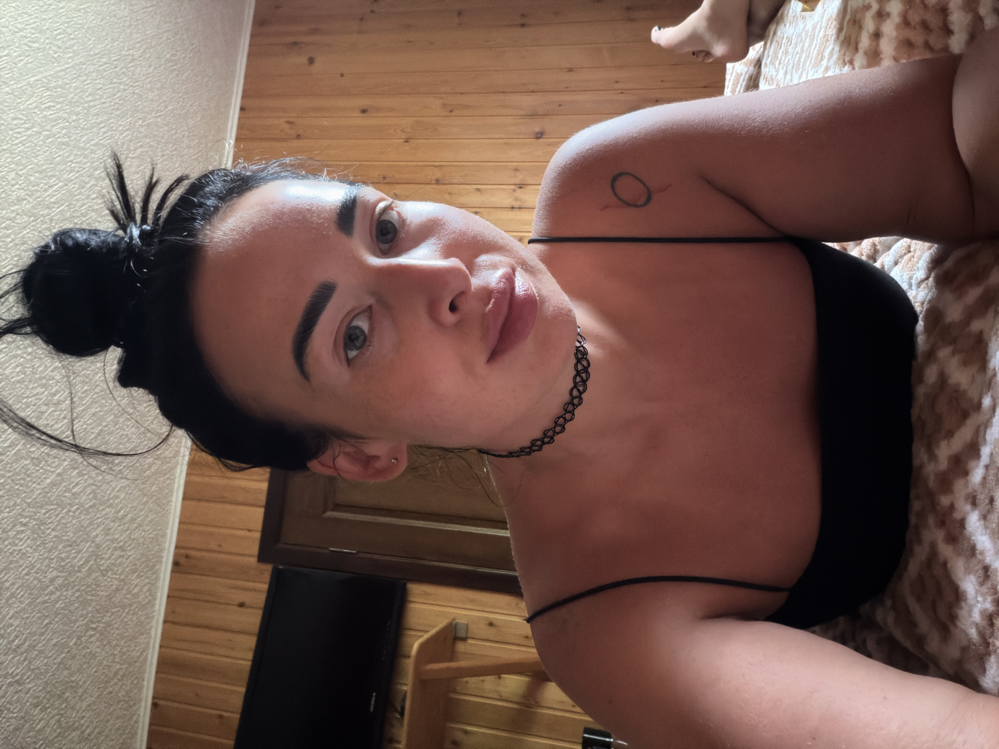 A woman is lying on a bed in a room with wood-paneled walls. She has her hair pulled up in a bun and is wearing a black spaghetti strap top and a black choker necklace. There is a small tattoo of a circle with a line through it on her right shoulder. A television is mounted on the wall to the left of the frame. The room appears to be in a hotel or guesthouse, possibly in Sochi, Russia.