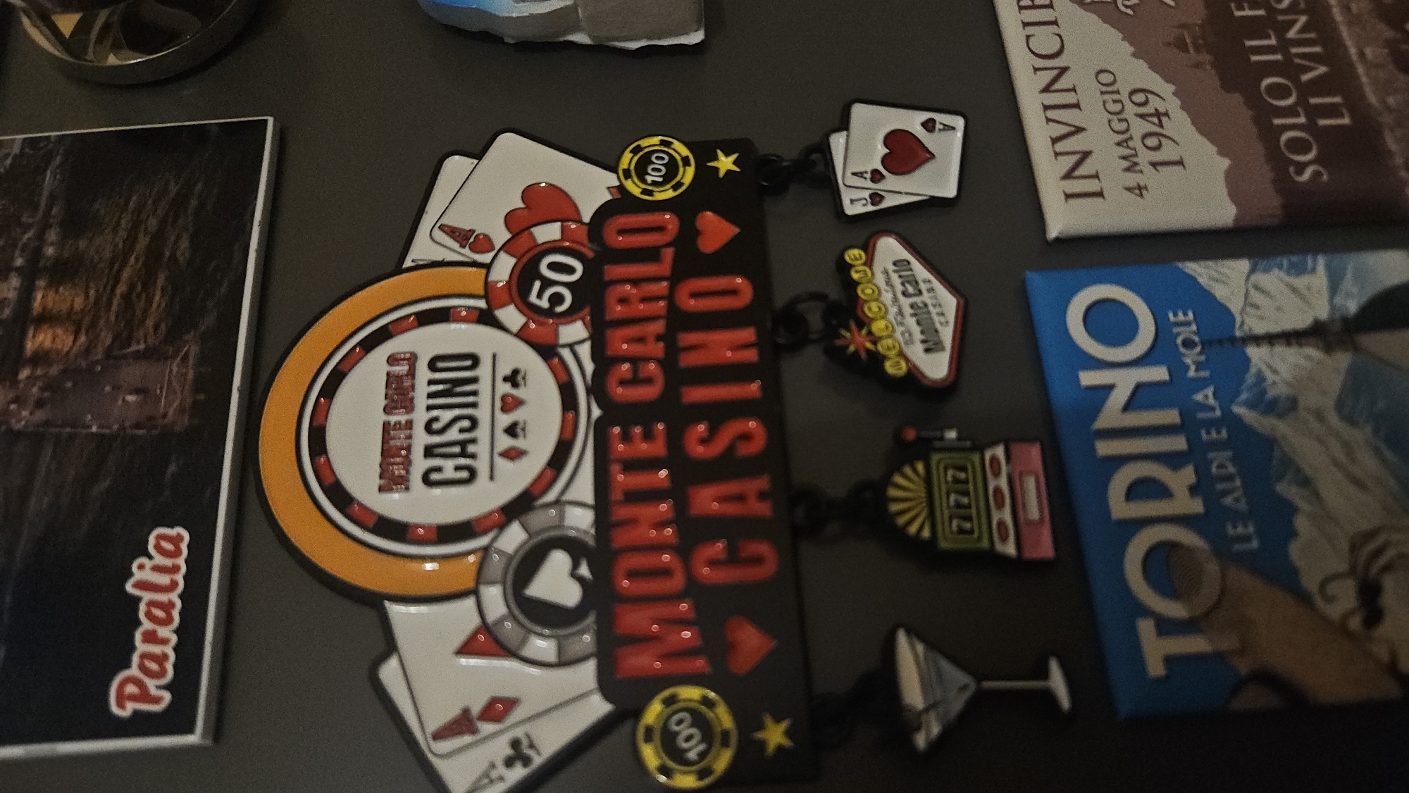The image displays an assortment of magnets on what appears to be a refrigerator door or a similar metallic surface. The magnets predominantly relate to travel and casino themes.
There's a magnet with the text "Paralia", another with "Monte Carlo Casino" and casino chips, playing cards, and other casino related objects. There is also a magnet that reads "Torino le aldi e la mole" and another magnet that reads "INVINCIBILE 4 maggio 1949 SOLO IL F LI VINS". The composition gives the impression of a collection accumulated from different places, possibly souvenirs. There's no information that suggests this image was taken in Plovdiv, Bulgaria or related to the city in any manner.