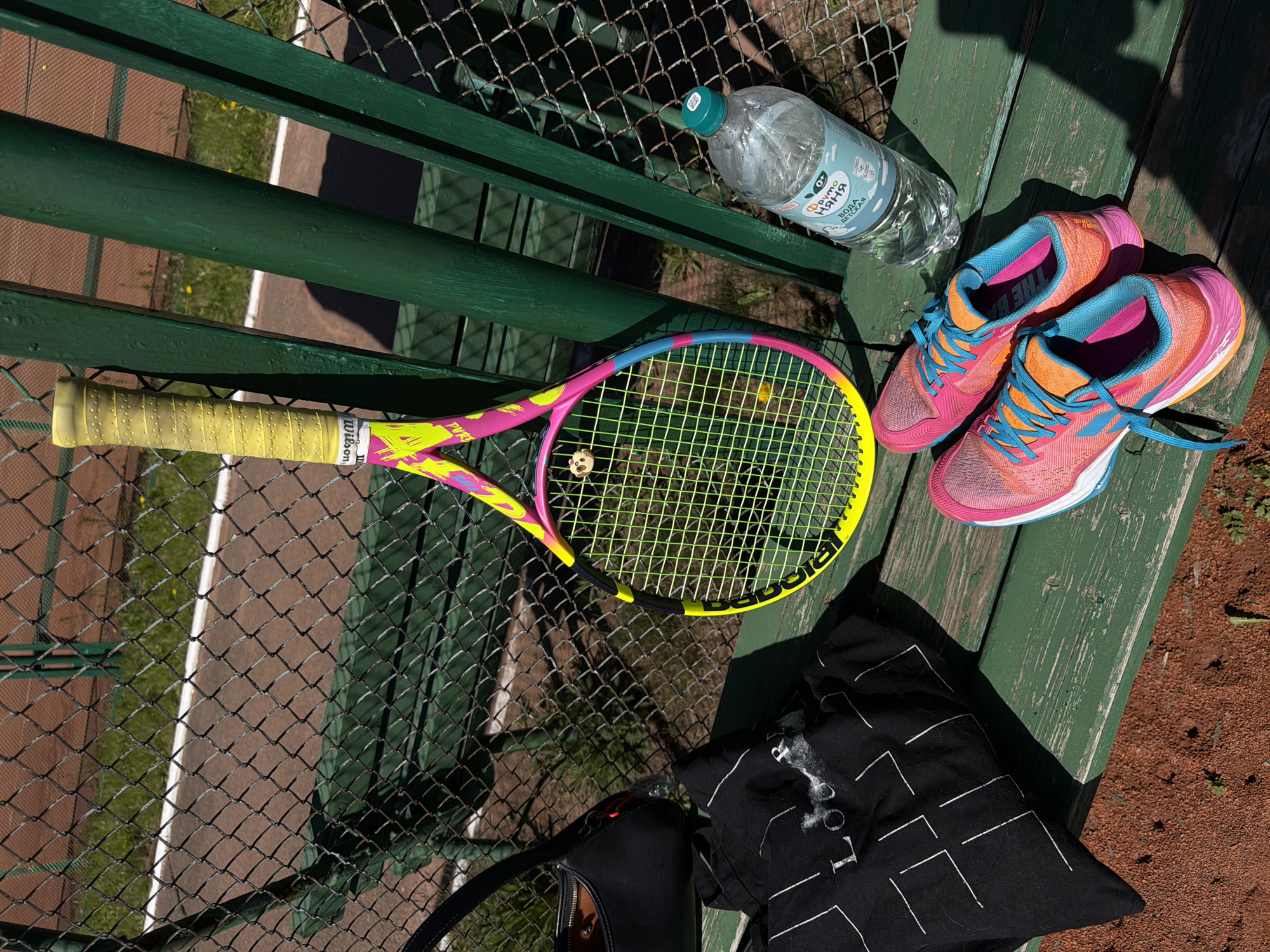 The image captures a vibrant collection of tennis equipment and personal items resting on a weathered green wooden bench, set against the backdrop of a tennis court. The bench, made of painted planks, shows signs of age and use.
**Subjects:** Prominently featured are a brightly colored tennis racket, a pair of athletic shoes, a plastic water bottle, a black handbag, and a black t-shirt. The tennis racket is pink and yellow with a bright yellow grip, lying diagonally across the bench. The running shoes are a striking combination of pink, orange, and blue. A clear plastic water bottle is positioned near the shoes. A black shoulder bag and a black t-shirt with white abstract patterns are also on the bench.
**Scene and Setting:** The bench is positioned adjacent to a chain-link fence, which offers a partial view of a clay tennis court with a white boundary line. Beyond the court, patches of green grass are visible. The scene is outdoors, likely at a sports facility or park in Krasnoyarsk, Russia.
**Activity or Event:** The presence of sports gear strongly suggests a tennis activity, either a break during a match or practice, or the equipment being laid out before or after play. The items appear to be temporarily set aside.
**Notable Details:** The time of day is marked by bright sunlight, casting distinct, sharp shadows from the fence and the objects, indicating a clear, sunny day, probably midday or early afternoon. The tennis court's red clay surface is a clear indicator of the sport.
**Visible Text:**
* On the water bottle, Russian text "Фруто НЯНЯ" (Fruto Nyanya) and "вода детская" (children's water) along with "0.33Л" are visible.
* The tennis racket grip features a "Wilson" logo, and "Babolat" is stenciled on the strings.
* Partially visible text on the tongue of one shoe appears to start with "THE B".
* The black t-shirt displays abstract white line patterns and the letters "LO".