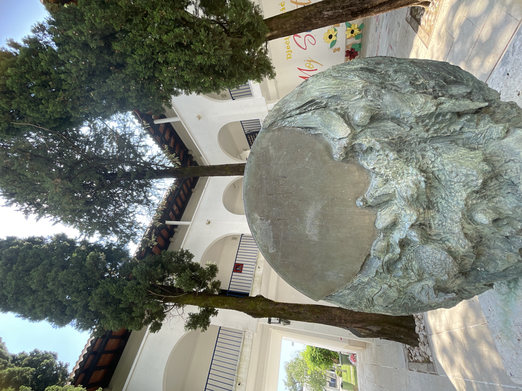 The image captures a large, weathered bronze sculpture of a human head situated in an outdoor space within the city of Budva, Montenegro. The sculpture’s face is relatively smooth and featureless, contrasting with the rough, textured mass forming the back of the head. The sculpture stands in front of a two-story white building with arched openings and blue railings. The building displays a sign with the text "Dječji Moj" above some illustrations, including sunflower decorations. The view includes trees that grow up between the building's facade and the statue. The ground is paved with light-colored stones, and sunlight is visible in the background, suggesting a clear day. No people are visible in the immediate foreground. The environment suggests a public or institutional setting, possibly a school or community center.