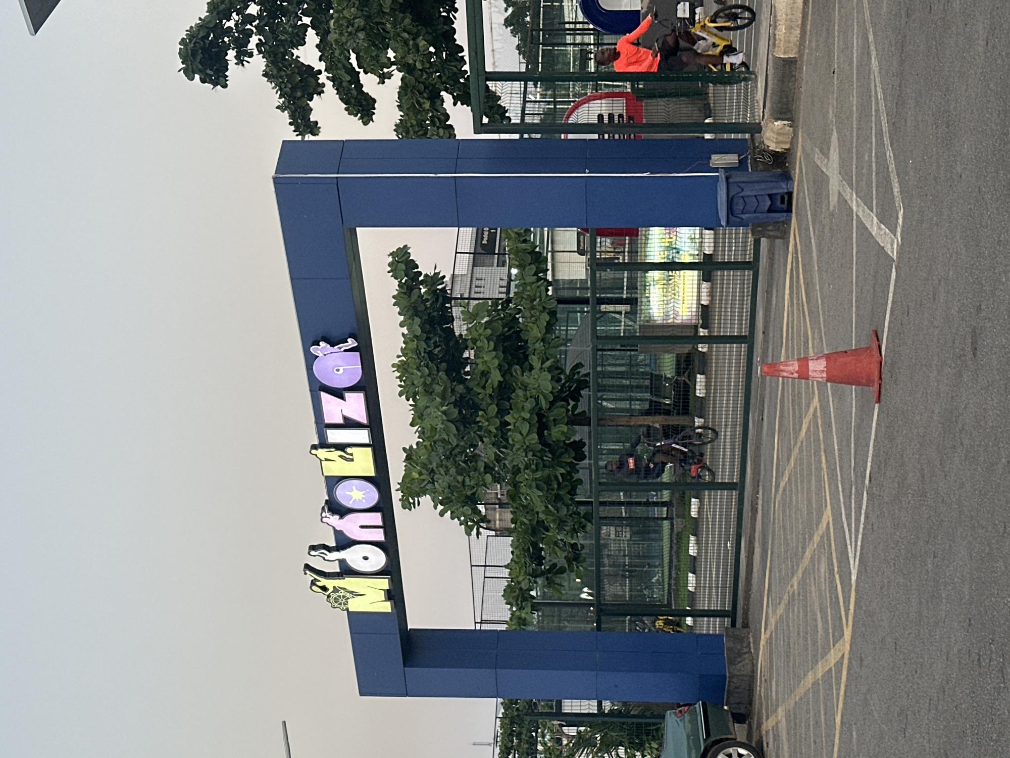 The image shows the entrance to a recreational facility in Abuja, Nigeria, with the name "Monkliza" displayed in illuminated letters above a blue archway. The letters are in various colors: yellow, white, pink, and purple, with stylized figures integrated into the design. On the left side of the arch, a green car is parked. A traffic cone is placed on the asphalt in the foreground. Behind the gate, there are tennis or padel courts with green fencing. A person in an orange shirt is riding a yellow bicycle on the right side of the image. In the background, several trees and buildings are visible under an overcast sky. The ground is paved with asphalt and marked with yellow and white lines, indicating a parking area.