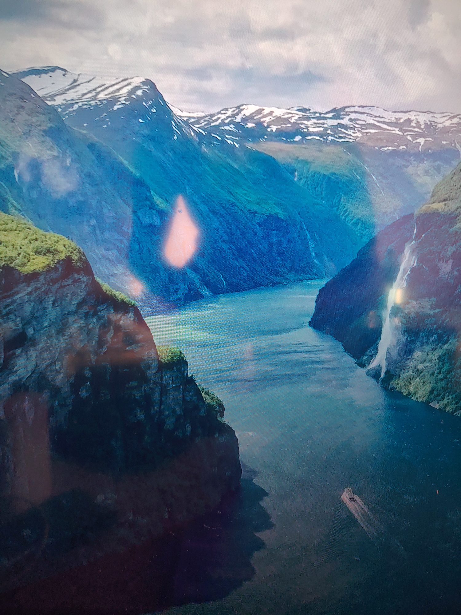 The image depicts a fjord, a narrow inlet of the sea between steep cliffs, with snow-capped mountains in the background. The water is a dark blue, and a boat is visible in the distance. The cliffs are covered in green vegetation in places. The location is likely Norway, known for its fjords, but the specific location within Norway is not identifiable from the image.