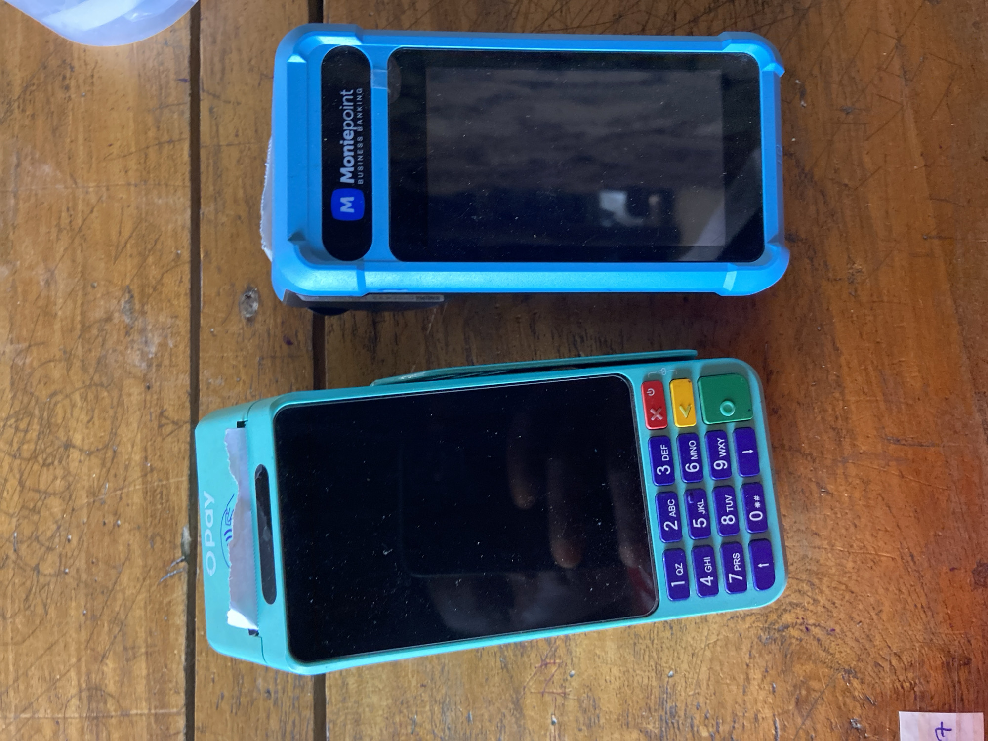 The image shows two point-of-sale (POS) terminals placed on a wooden surface. The top terminal is turquoise and has a keypad with numbers and letters, a screen, and the logo "Opay" on its side. The bottom terminal is blue and has a screen with the logo "Moniepoint BUSINESS BANKING" on its side. There is a small piece of paper with writing on it to the left of the turquoise POS terminal. The wooden surface appears to be a table or counter. The scene is indoors.