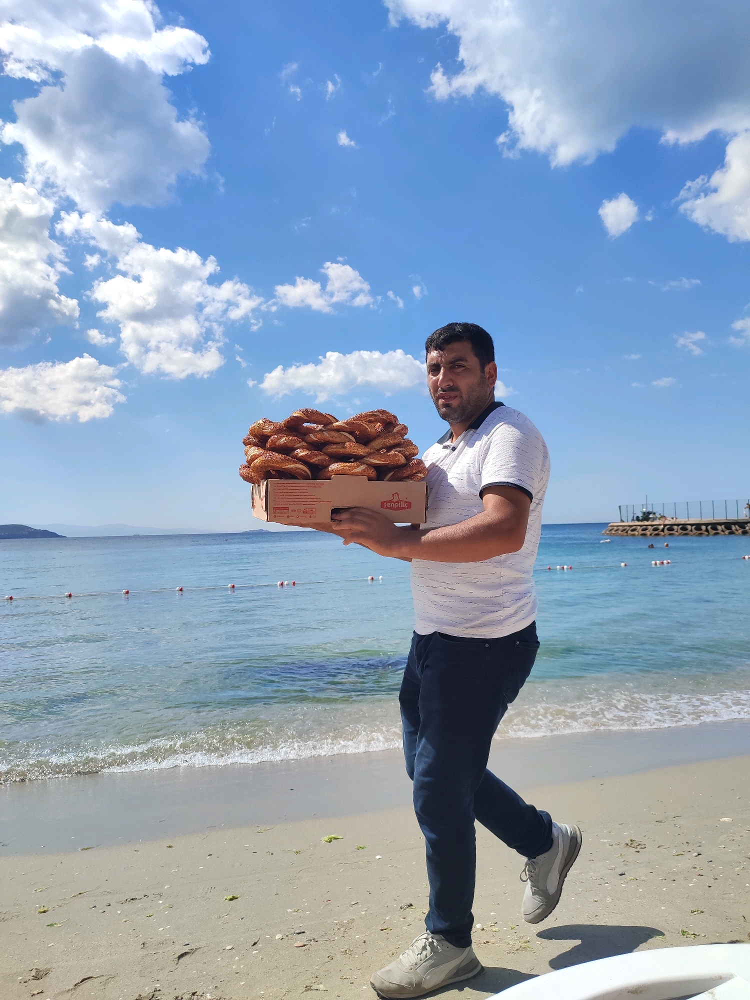 A man stands on a sandy beach, holding a cardboard box filled with simit, a Turkish sesame seed bread. The sea is to his right, with calm waves and buoys marking a swimming area. In the background, a pier with structures and people can be seen. The sky above is bright blue with fluffy white clouds. The scene appears to be a sunny day, likely in a coastal area of Kadıköy, Turkey, known for its beaches and seaside offerings. The man is wearing a white t-shirt, dark jeans, and athletic shoes, and he is looking towards the camera. The box of simit has some text and a logo on its side.