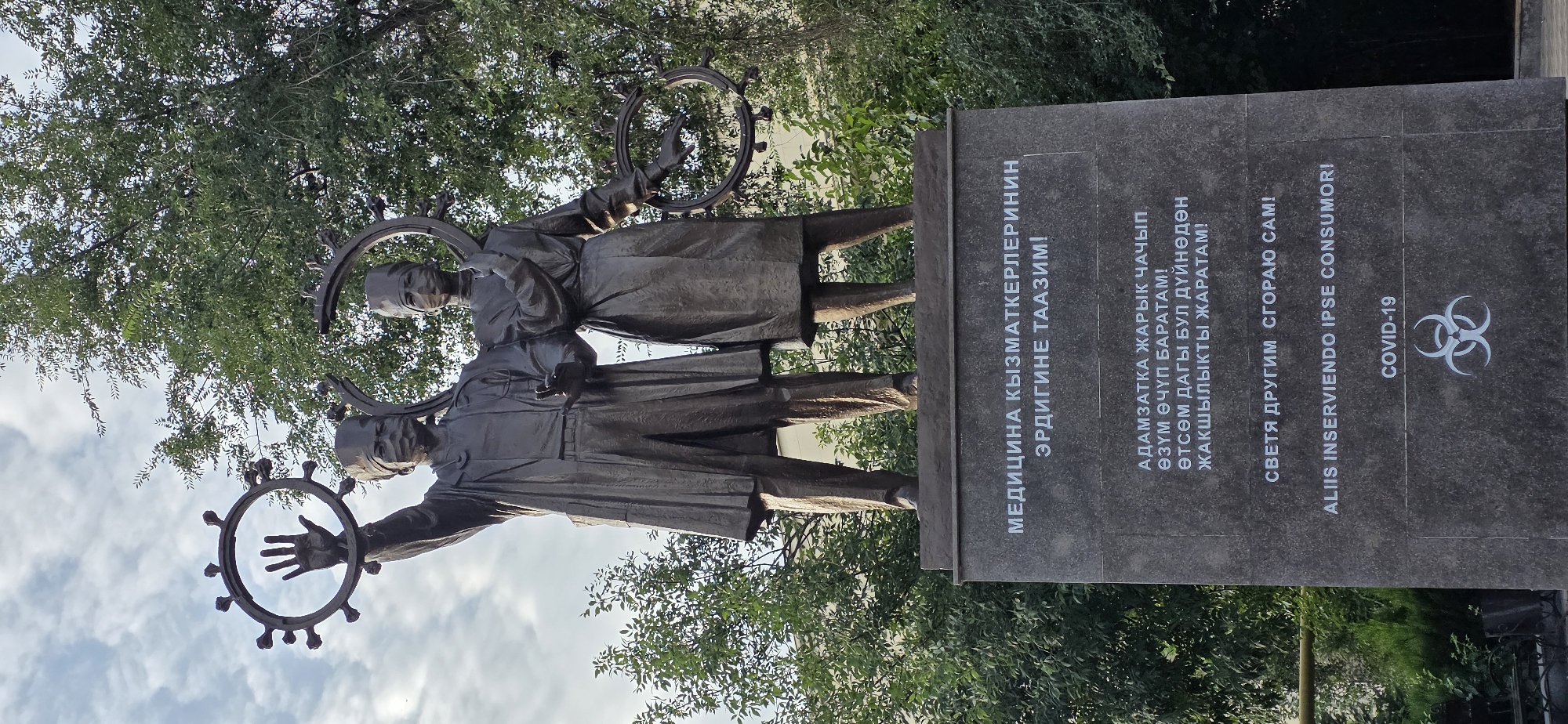The image shows a bronze statue of two medical professionals, a man and a woman, standing on a granite pedestal. Both are wearing masks and medical attire. The man on the left is holding a ring-like object with a hand symbol in the center, raised above his head. The woman on the right is holding a similar ring. The pedestal is inscribed with text in multiple languages, including Kazakh, Russian, and Latin. The Kazakh text reads "МЕДИЦИНА ҚЫЗМАТКЕРЛЕРІНІҢ ЭРДІГІНЕ ТААЗІМ!", which translates to "Homage to the Valour of Healthcare Workers!". Below this, in Kazakh, it says "АДАМЗАТҚА ЖАРЫҚ ЧАЦЫП ӨЗУМ ӨЧУП БАРАТАМ!ӨТСӨМ ДАҒЫ БУЛ ДҮЙНӨДӨН ЖАҚШЫЛЫҚТЫ ЖАРАТАМ!". In Russian, it reads "СВЕТЯ ДРУГИМ СГОРАЮ САМ!", meaning "Burning myself to light up others!". The Latin inscription states "ALIIS INSERVIENDO IPSE CONSUMOR!", which also translates to "By serving others, I consume myself." At the bottom of the pedestal, "COVID-19" is written, accompanied by a biohazard symbol. The statue is located in Bishkek, Kyrgyzstan, and is a tribute to healthcare workers during the COVID-19 pandemic. The background consists of green trees and a cloudy sky.