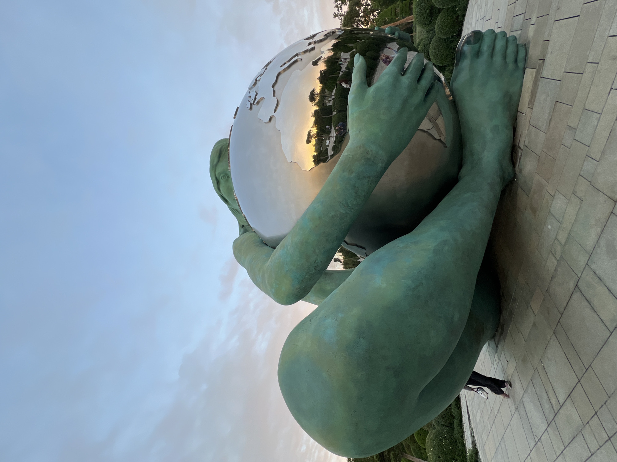 This striking image captures a large, green-patinated bronze sculpture set outdoors in Krasnodar, Russia. The central figure, appearing to be female, is depicted in a kneeling or seated position, embracing and cradling a massive, highly polished, spherical object. This sphere resembles a globe or the Earth, its metallic surface reflecting the surrounding environment.
Reflected vividly in the sphere are elements of the scene: a warm, golden-orange sky indicative of sunset or sunrise, dark silhouettes of trees and manicured hedges, and what appears to be part of a structure or building. The smooth, reflective surface of the globe sharply contrasts with the textured, weathered appearance of the bronze figure, which shows varying shades of green, blue, and brown patina.
The sculpture rests on a paved surface, made of rectangular tiles in a muted grey tone. In the bottom right corner, a person's lower legs and feet, clad in dark trousers and light-colored shoes, are visible near the sculpture's base, providing a sense of the artwork's substantial scale. The background features a partially cloudy sky with soft, diffused light, further suggesting either early morning or late afternoon. The overall impression is one of guardianship or a deep connection to the Earth, with the figure's posture conveying a sense of embrace or burden. No visible text is present in the image.