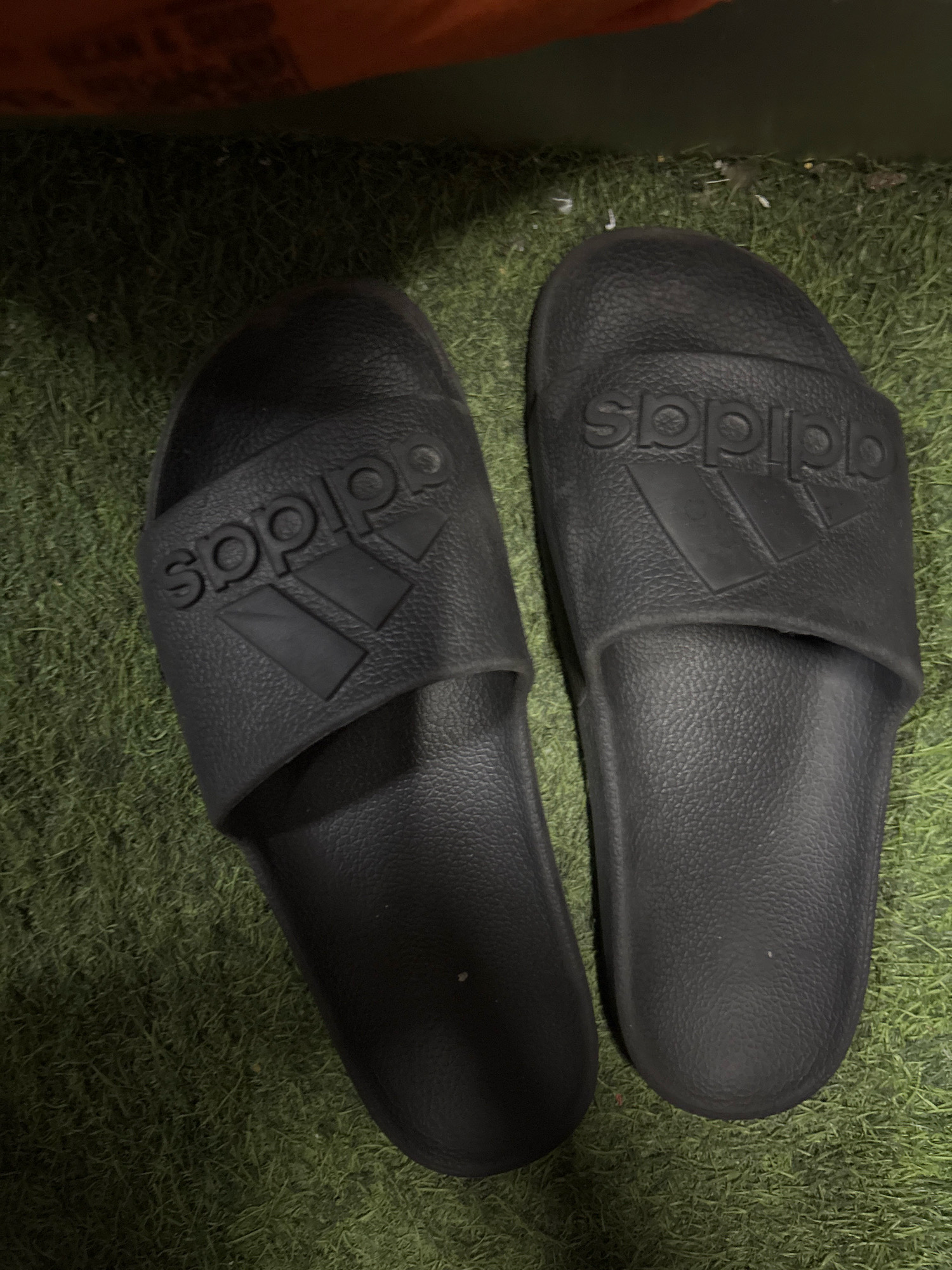 Two black Adidas slides are positioned on a patch of artificial green grass. The slides are placed side-by-side, with the left slide slightly angled and the right slide more directly facing the viewer. The Adidas logo, featuring three parallel stripes and the brand name, is embossed on the strap of each slide. The background is blurred, showing a darker area above the grass, suggesting an indoor or shaded outdoor environment. There are no people or visible actions in the image. The scene is static and focuses solely on the footwear.