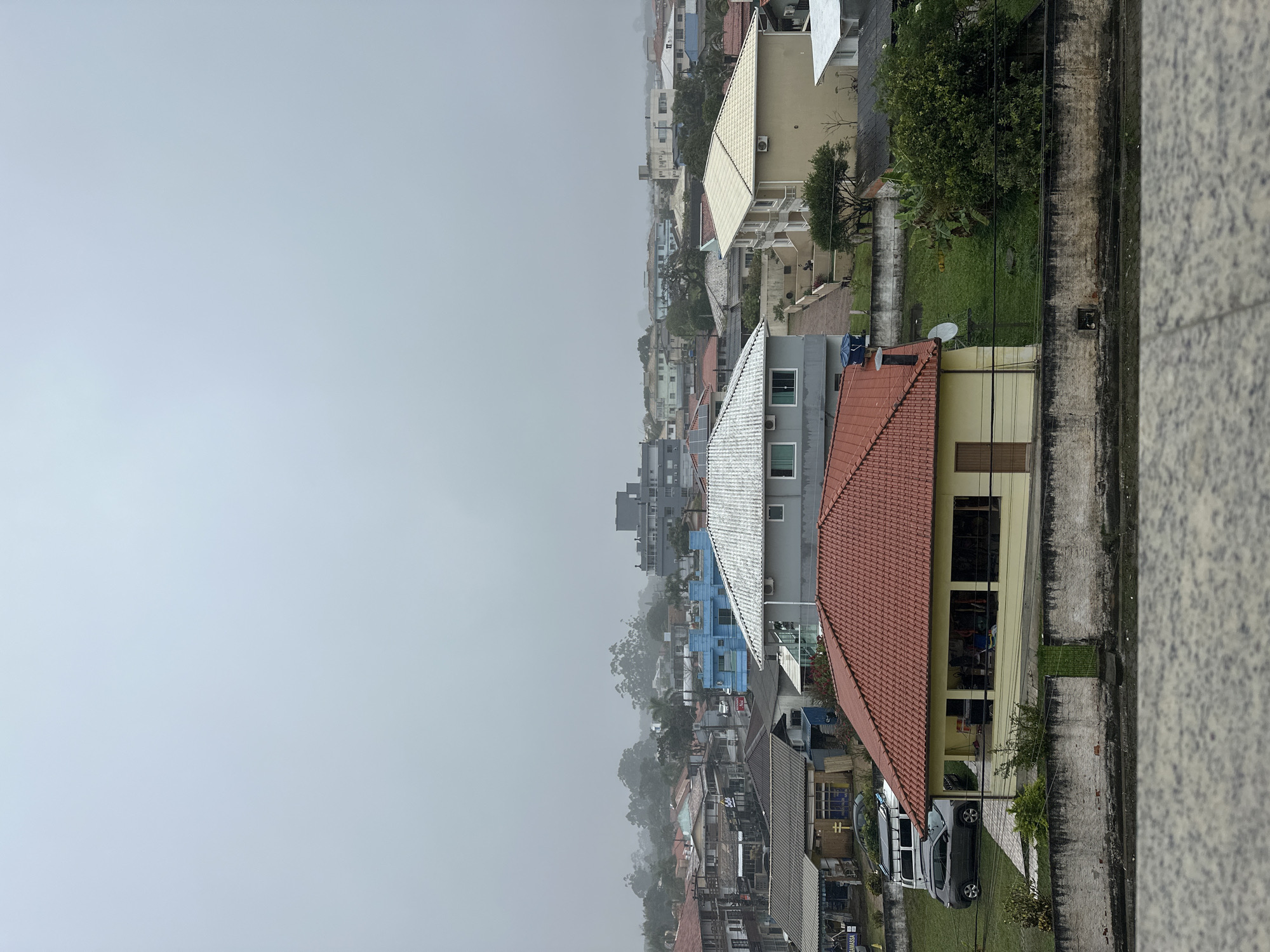 The image shows a residential area in Florianópolis, Brazil, under a hazy, overcast sky. The view looks out over several houses with tiled roofs, mostly in shades of red and yellow, interspersed with some modern apartment buildings. A few cars are visible, including a dark gray SUV and a white van. Green lawns and trees surround the houses. The foreground shows a textured concrete surface, likely a rooftop or balcony. The overall atmosphere is subdued due to the lack of direct sunlight.
