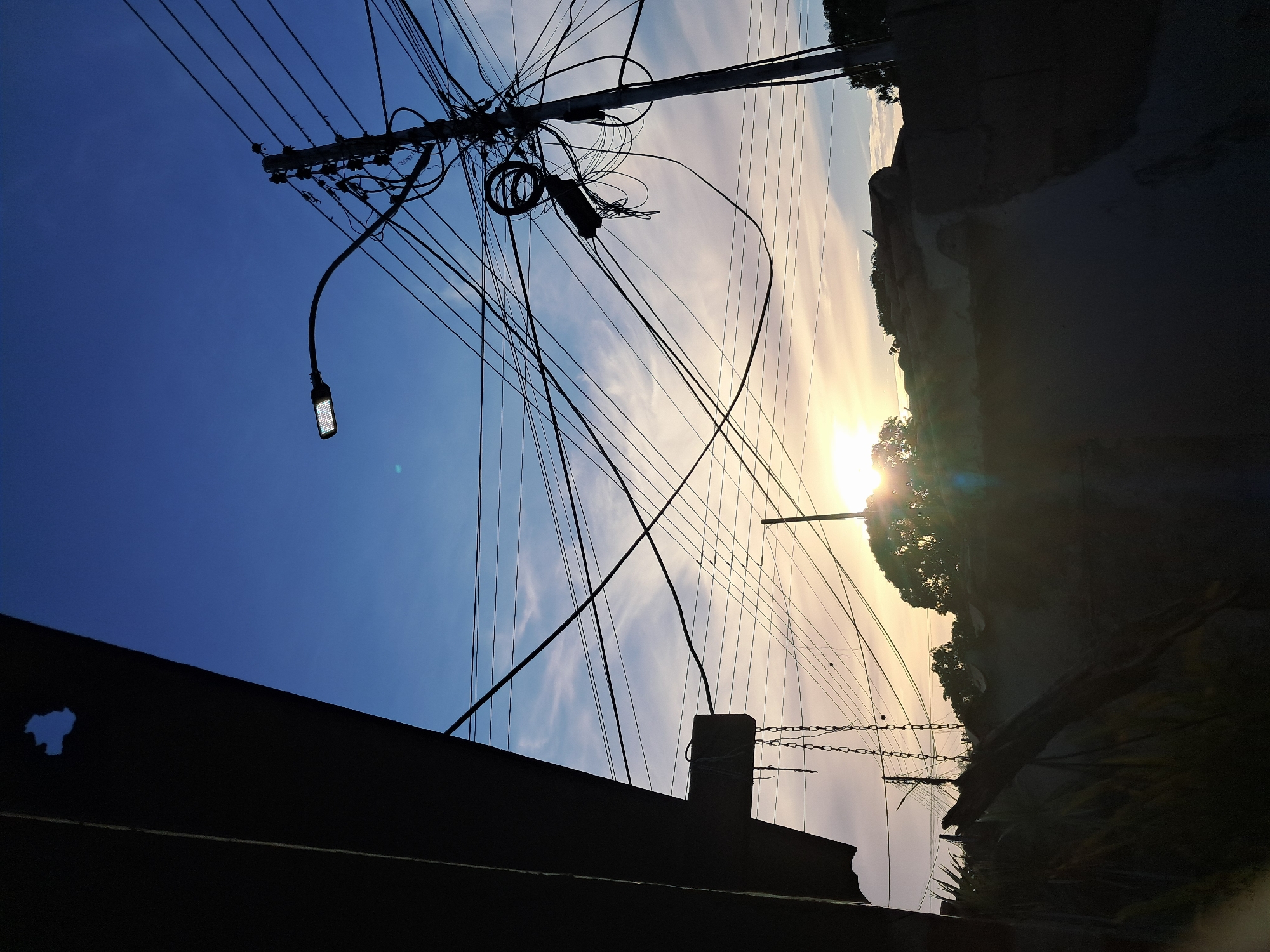 The image depicts a view in Juan Griego, Venezuela, characterized by a cluttered urban landscape. The sun is low on the horizon, casting a bright glare that obscures details. Overhead, a complex network of power lines and cables crisscrosses the sky, connected to a utility pole equipped with a street light. Silhouetted against the sky are rooftops and vegetation, indicating a residential area. In the foreground, the dark shape of a building structure adds depth to the scene.