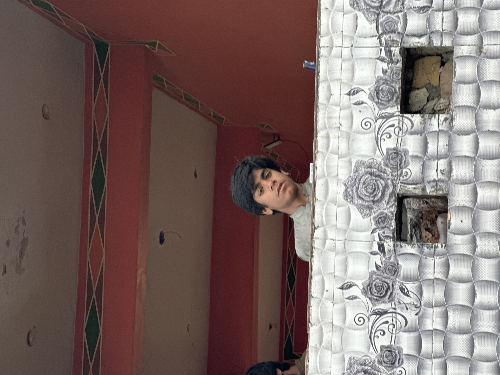 The image depicts a residential interior with two individuals present. A young man with dark hair peers from behind a tiled wall that features floral patterns and two square openings exposing brick and rubble. The interior space behind the wall is defined by reddish-brown walls and patterned trim, with visible electrical wiring. A second individual is partially visible at the bottom center of the frame. The location is Unknown, Unknown.