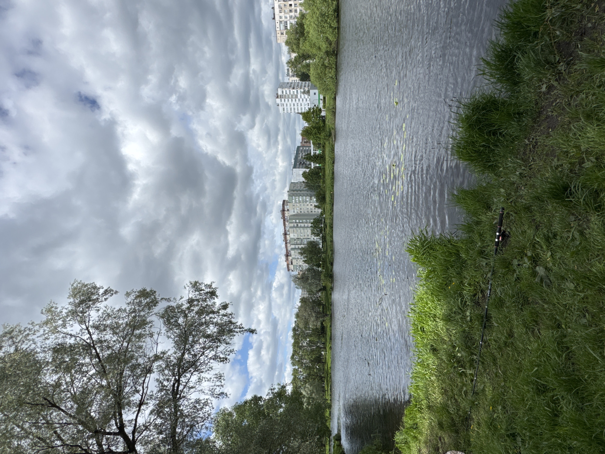 The image shows a body of water, likely a canal or river, with buildings visible in the background. The sky is overcast with heavy clouds. A fishing rod is leaning against the grassy bank on the right side of the image. Trees line the waterway. The buildings in the background suggest a residential area. It could be in Saint Petersburg, Russia. The setting is an urban natural environment.
