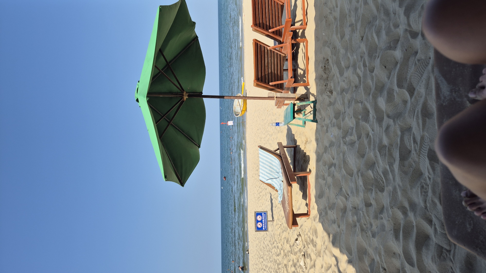 This is a photograph of a beach scene in Da Nang, Vietnam. In the foreground, the sandy beach is covered in footprints and the bottom portion of a person's legs are visible, suggesting they are lying on a lounge chair. Further back, a green beach umbrella provides shade over two wooden lounge chairs. To the left of the umbrella, another wooden lounge chair is set up with a blue and white striped towel draped over it. A small, green table sits in front of this chair, with a plastic water bottle on top. To the left of the lounge chairs, a blue sign with white symbols is posted in the sand. The ocean stretches out in the background under a clear blue sky, with a few people visible in the water. Several boats are scattered on the water, including a yellow one near the shore. The overall impression is of a relaxing day at the beach.