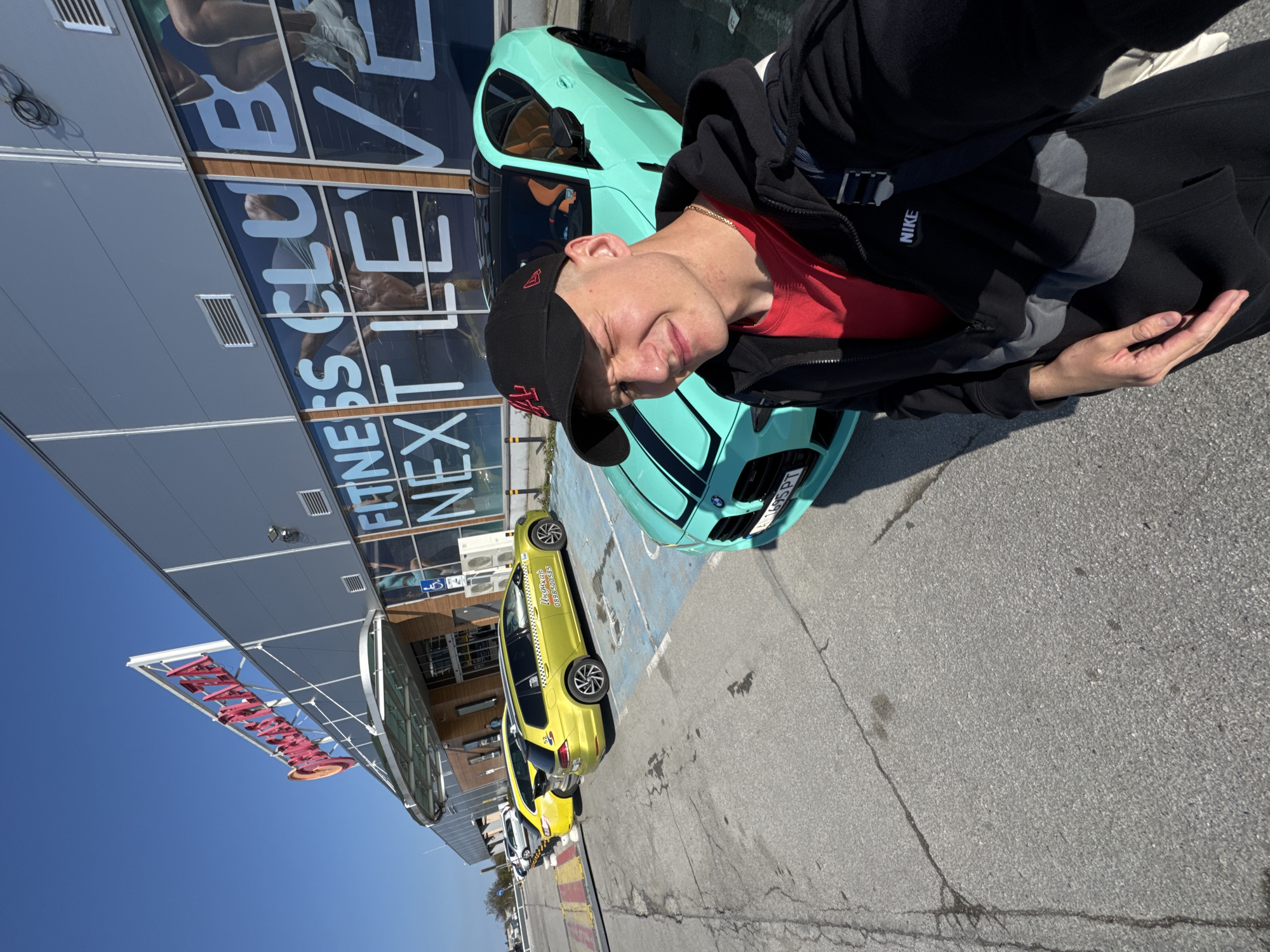 The image captures a man posing in front of a light green car in an outdoor parking lot. He wears a red shirt, black jacket, and a black baseball cap with red lettering. The car behind him is light green with a black stripe.
Behind the car is a building with reflective windows that read "Fitness Club Next Level." To the left of the building is a glass-roofed entrance with a red sign above. Further to the left are several yellow taxi cabs lined up.
The image appears to be taken in Burgas, Bulgaria based on the writing on the taxi cabs. The weather is sunny and the sky is clear.