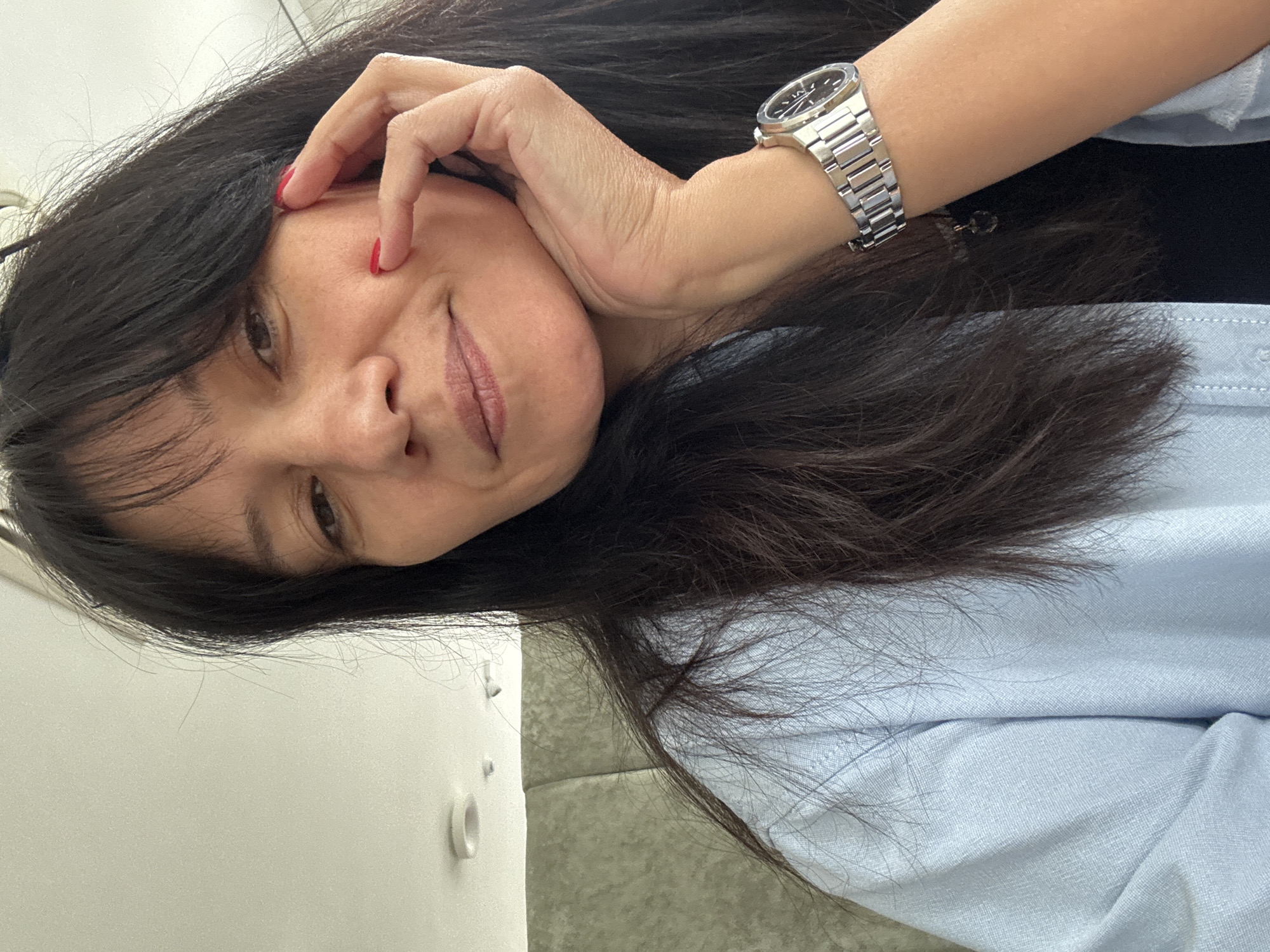 A woman is posing for a selfie in an indoor environment. She is wearing a light blue shirt and a silver watch on her wrist. Her hair is dark and long. Her fingers are touching her face, and her nails appear to be painted red. The background is out of focus but appears to be a wall. The city and location are unknown.