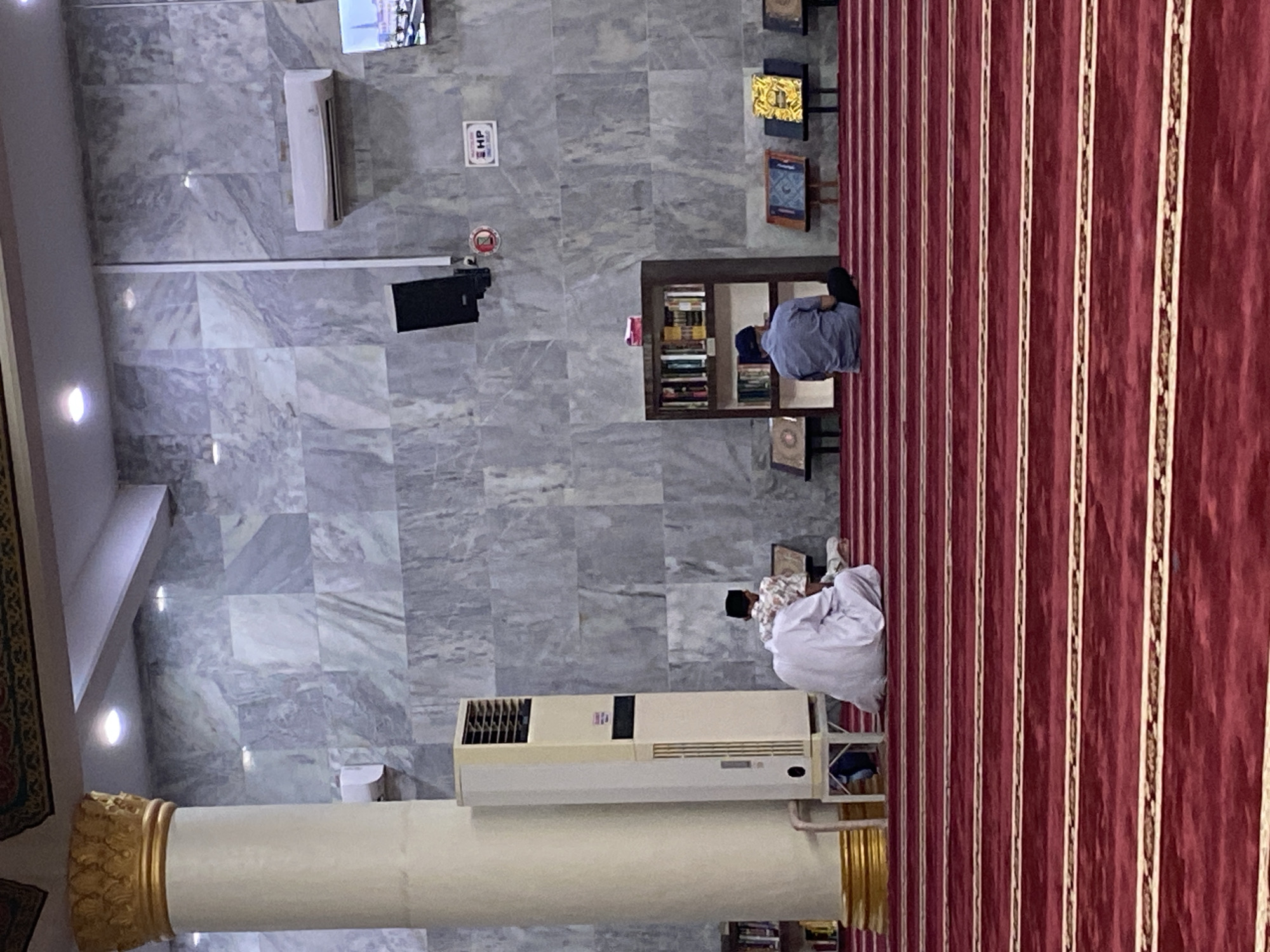 The image depicts the interior of a mosque, specifically showing the prayer area with a patterned red carpet. In the foreground, the carpet features prominent parallel gold and white stripes. Two men are visible in the mid-ground, engaged in prayer or reading. One man, wearing a white robe and a cap, is seated on the carpet facing a large, vertical, cream-colored air conditioning unit. Another man, dressed in a blue shirt and dark pants, is seated on the carpet facing away from the camera, towards a bookshelf.
The mosque's interior features marble-patterned walls. To the left, a large white pillar with a gold capital stands prominently. To the right of the pillar, a tall, vertical air conditioning unit is situated. Mounted on the wall above the bookshelf is a black speaker. Further to the right, a large flat-screen television displays an image, possibly of a city skyline. A small sign with the letters "HP" is also visible on the wall. A bookshelf filled with books is positioned against the wall. Several books are also displayed on stands in front of the bookshelf. The ceiling has decorative moldings and recessed lighting. The overall environment suggests a place of worship and quiet contemplation. This mosque is located in Lhokseumawe, Indonesia.